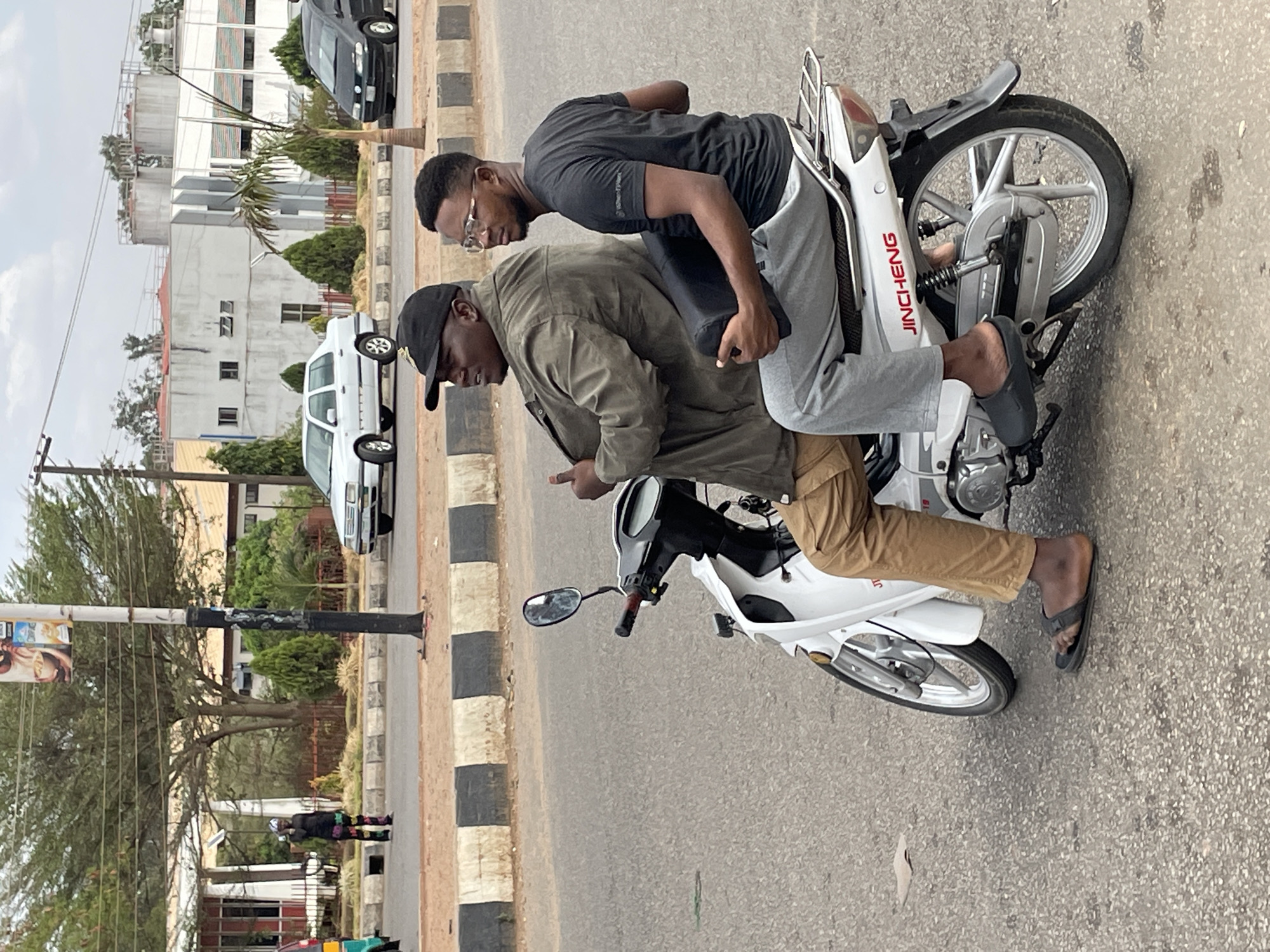 Two men ride a white Jincheng brand motorcycle along a paved road with black and white striped curbing, likely in Jos, Nigeria. The driver, wearing a khaki jacket and pants and a black cap, points with his left hand. The passenger, wearing a black shirt and grey pants, sits behind him. Both men wear sandals. In the background are buildings, vehicles, trees, and a utility pole with advertisements. The sky is overcast.