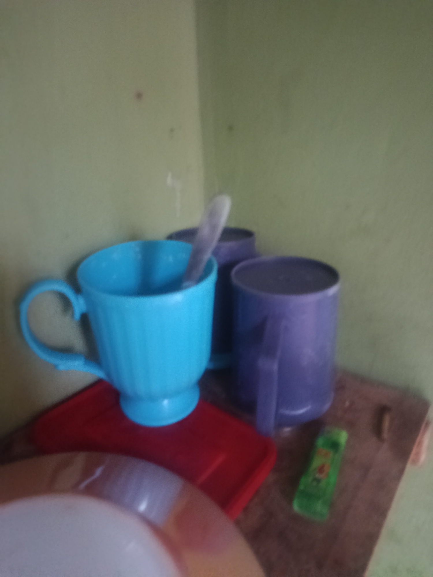 In a dimly lit room with light green walls, three cups and a spoon are placed on a wooden surface. A bright blue cup with a textured exterior and a matching handle sits on a red tray. Inside the blue cup, a silver spoon is partially visible. To the right of the blue cup, two purple cups are positioned. The first purple cup is slightly taller and appears to have a wider opening. The second purple cup, positioned behind the first, is shorter and appears to be a deeper shade of purple. A small green object with red and yellow markings rests on the wooden surface between the cups and the viewer's perspective. The scene appears to be a still life, with no people or discernible action. The overall atmosphere is quiet and domestic.