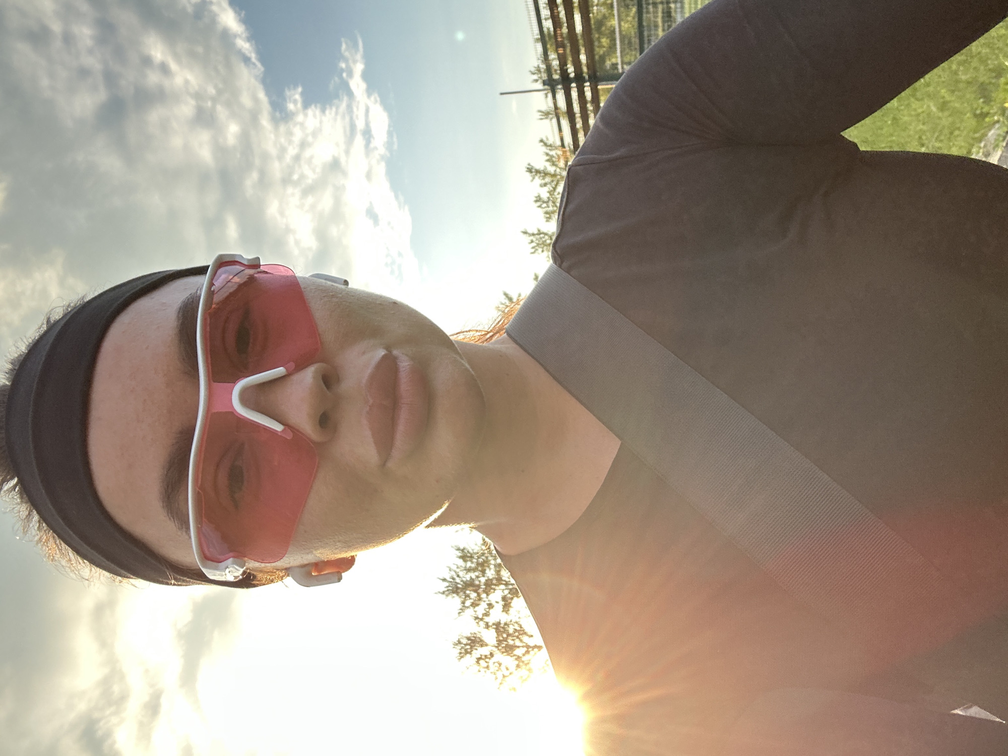 A close-up selfie captures a woman wearing a black headband, pink athletic sunglasses, and white earbuds. She has on a dark-colored long-sleeved shirt with a grey strap across her chest. The background consists of a bright sky with clouds, trees, and a fence. The sun is setting or rising, casting a warm glow on the scene, with lens flare visible on the left side of the image. The location is in the city of Yuzhnouralsk, Russia.