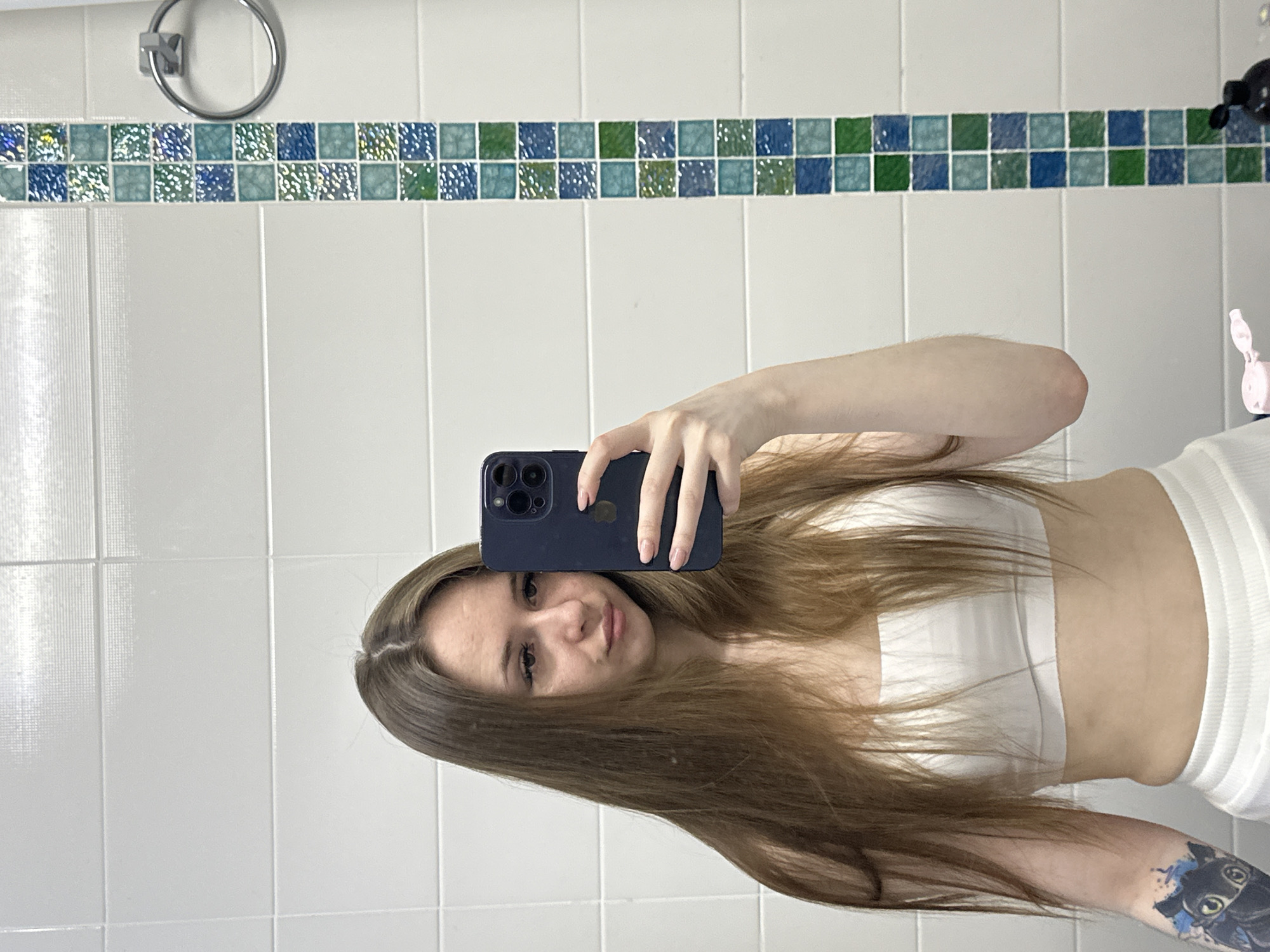 An image, rotated 90 degrees counter-clockwise, depicts a young woman taking a mirror selfie in what appears to be a bathroom. The woman, with long, light brown hair parted in the middle, looks directly at the camera with a neutral to slightly smiling expression. She is wearing a white strapless top and white ribbed bottoms. Her left arm, extended to hold a dark-colored smartphone with a visible camera array, reveals a distinctive tattoo on her forearm – a black cat-like creature, possibly Toothless, with blue paint-like splashes. Her fingernails are painted with a light pink or nude polish.
The background consists of white rectangular wall tiles. A horizontal band of smaller, square mosaic tiles in varying shades of blue, green, and teal runs across the upper part of the wall. To the left, a chrome-plated circular towel ring is mounted on the white tiles. On the far right, a small pink object, possibly an electric toothbrush or a bottle cap, is partially visible. The overall setting suggests an indoor environment, likely a bathroom, given the fixtures and tile work. There is no visible text in the image.