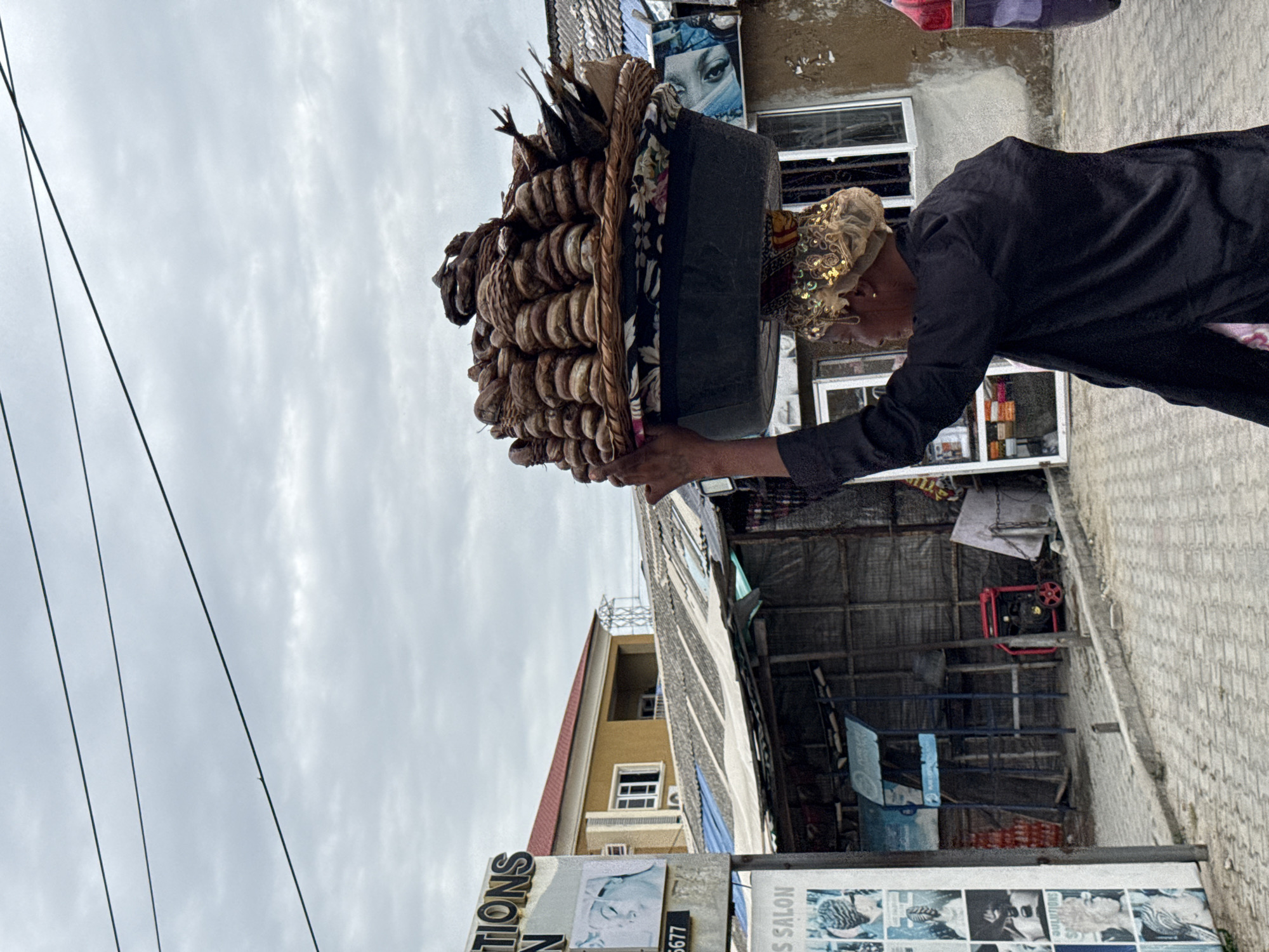 Ado, Nigeria: A woman in a long-sleeved black shirt carries a large, ornate basket filled with items (likely baked goods or similar foodstuffs) on her head. She is walking along a paved street. Behind her is a shop with visible advertising and a multi-story building. The sky is overcast.