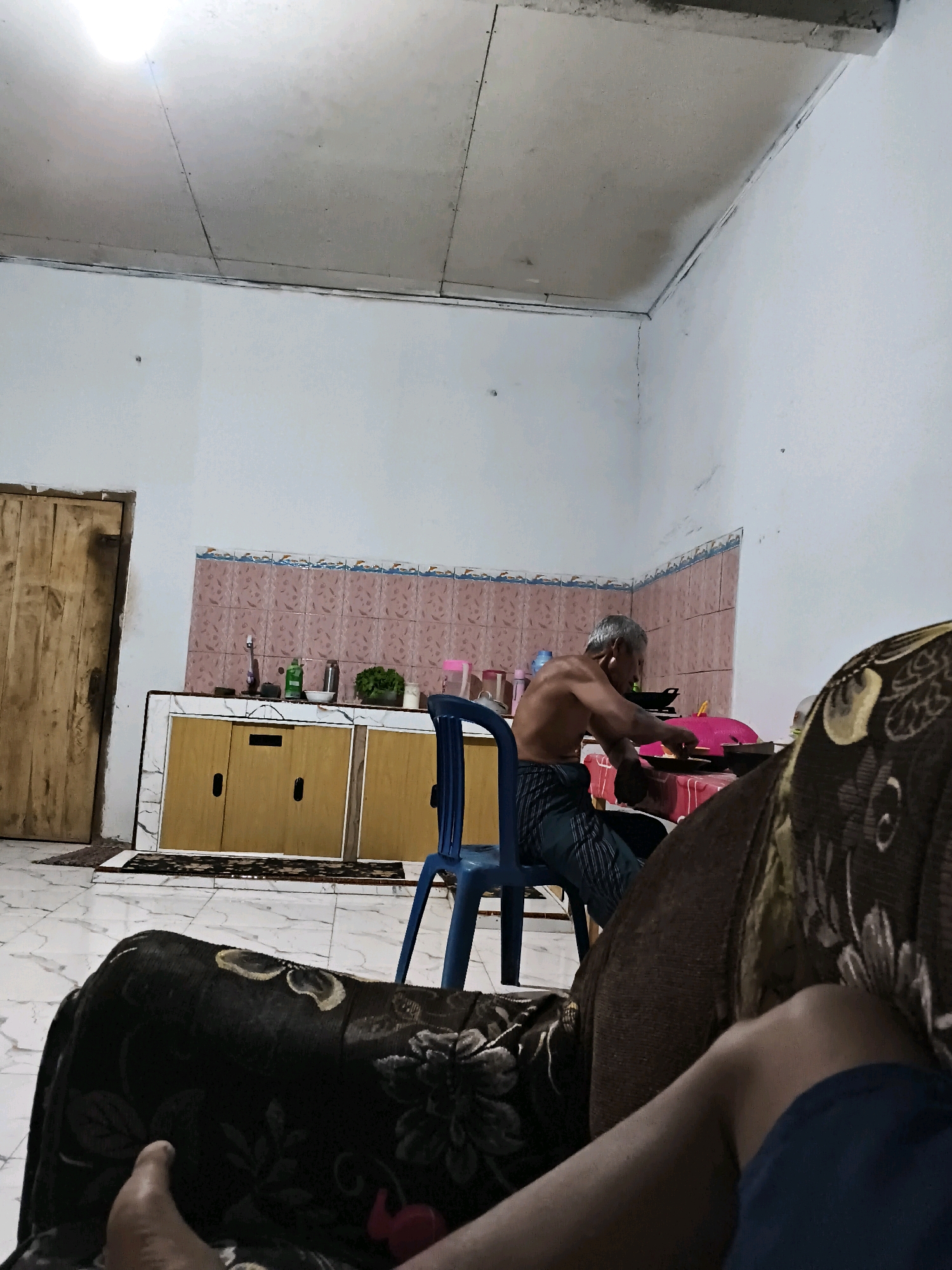 An elderly, shirtless man with graying hair is seated on a blue plastic chair in a room, facing away from the camera. He is wearing striped pants and appears to be engaged in an activity at a table covered with a red tablecloth, possibly preparing food. In the background, a kitchen area is visible with pink tiled walls above light brown cabinets. A wooden door stands to the left of the cabinets. In the foreground, the back of a patterned sofa and a person's leg are visible, suggesting the photograph was taken from a seated position on another sofa. The room has white walls and a textured ceiling, with a light source illuminating the upper portion of the room. The floor is tiled with a marble pattern. The overall setting suggests a residential interior in Banyuwangi, Indonesia, likely a kitchen or dining area.