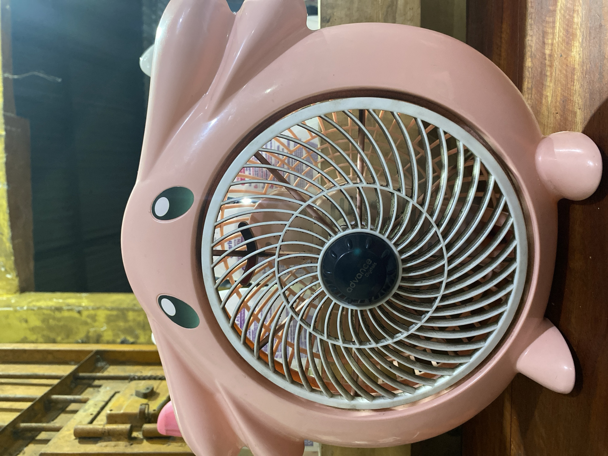 A pink, novelty fan shaped like a cartoon character sits on a wooden surface. The fan has large, cartoon-like eyes and ears. The center of the fan has a black circular control panel with the words "advance Digitals" printed on it. In the background, a yellow, textured wall is visible, along with a metal gate. The scene is dimly lit, suggesting it is indoors. The location is described as Ta', Indonesia.