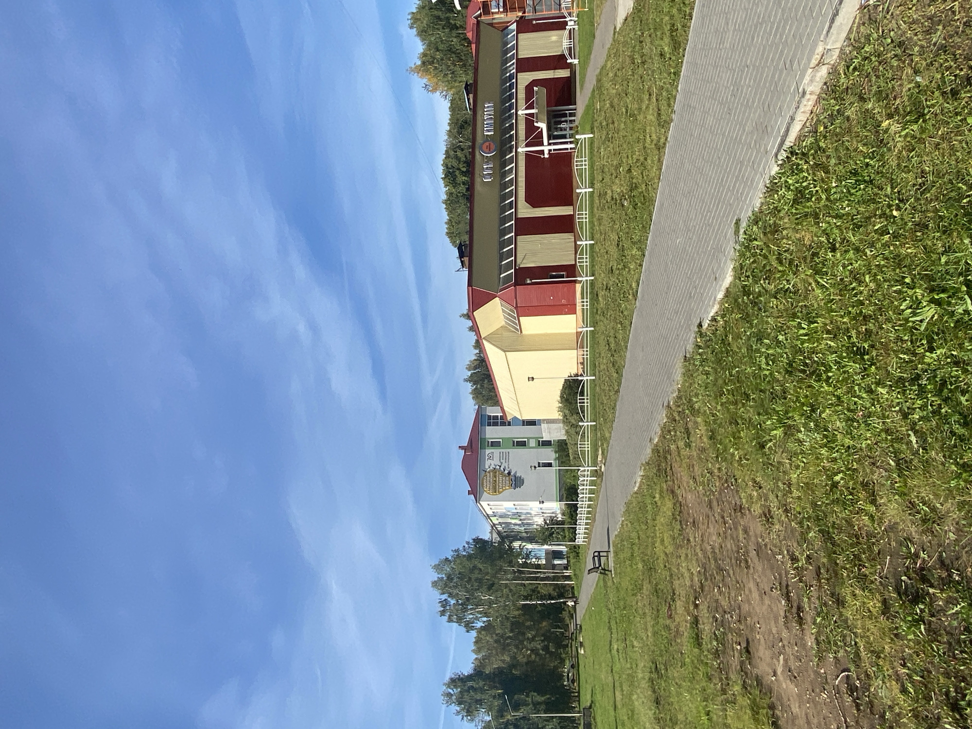 The image captures a sunny day in Chaikovsky, Russia, with a clear blue sky featuring wispy clouds. In the foreground, a paved walkway made of grey rectangular bricks leads into the distance. To the left of the walkway, a grassy area with some patches of bare earth slopes gently downwards, and a solitary park bench is visible. Further back, a line of white fences with decorative posts separates the walkway from a large building with a red roof and a facade predominantly in red and yellow. Above the entrance of this building, text is visible, including the word "КИМСТАЛ" with a logo. To the left of this building, another building with a light gray facade, green trim, and a red roof stands. It has several windows and a large mural depicting a book and text in yellow. A few trees with green foliage are scattered throughout the scene, with a taller cluster of trees behind the buildings. A lone pedestrian, a small figure in white, is walking on the grass in the distance to the left. The overall impression is of a well-maintained urban park or public space on a pleasant day.