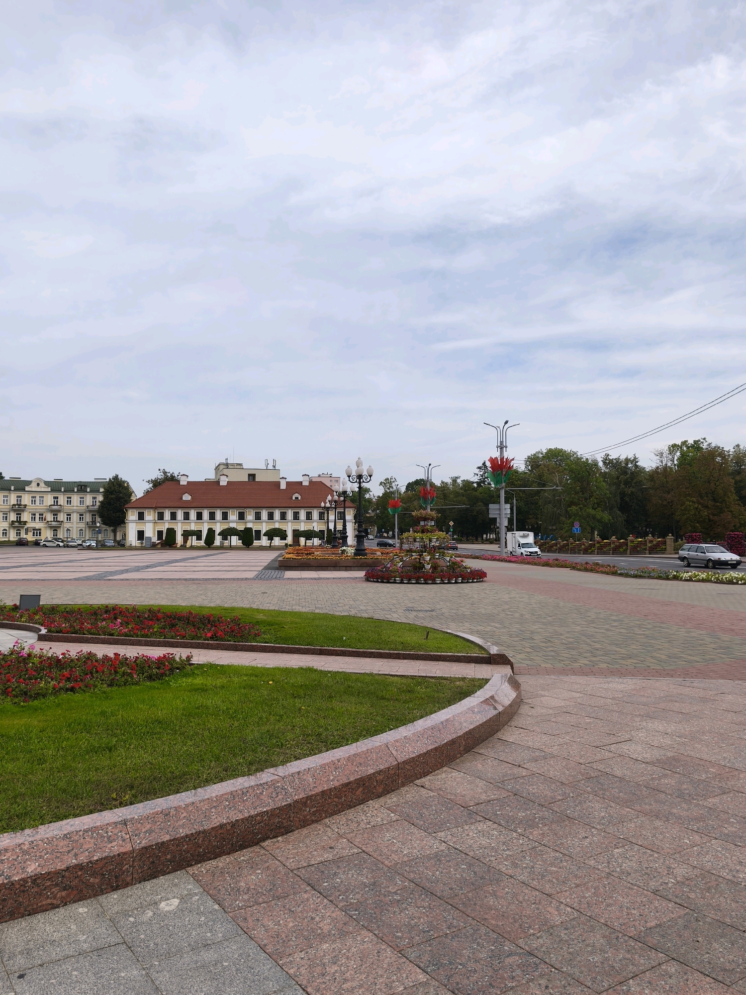 The image depicts a wide, paved plaza in Hrodna, Belarus, in front of a long, tan and white building with a red-tiled roof. The building appears to be an administrative or public structure, with multiple windows and a symmetrical facade. To the left of this building is a taller, light yellow building with a mansard roof, also featuring numerous windows. The plaza itself is paved with intricate patterns of light and dark stones, and features a raised flower bed with red and pink flowers in the foreground. Further into the plaza, there is a decorative fountain with multiple tiers, surrounded by more flower arrangements. Tall lamp posts with decorative elements are visible. Beyond the plaza, a road runs parallel to the buildings, with trees lining the far side. Cars are visible on the road, including a white van and a silver sedan. The sky is overcast with scattered clouds. The scene is peaceful and orderly, suggesting a well-maintained urban public space.