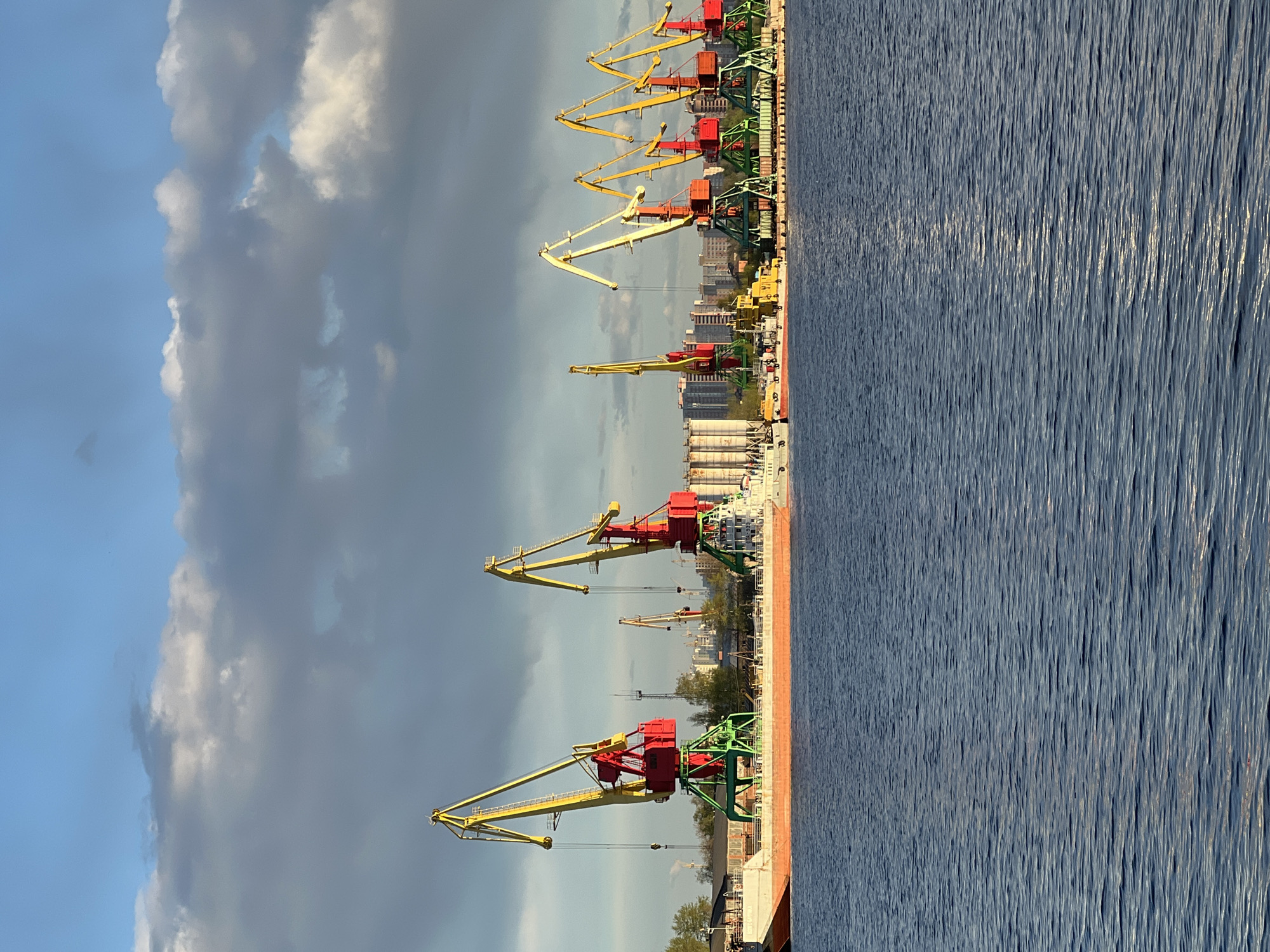 The image depicts a waterfront scene in what is presumably the city of Cherepovets, Russia, though specific identifiers confirming this location are not evident in the visual data. The view is dominated by a series of tall, industrial cranes positioned along the water's edge, likely within a port or shipping facility. The cranes are brightly colored with red cabs, yellow booms, and green support structures. A body of water, possibly a river or canal, is present, with visible surface texture indicating slight ripples or waves. Beyond the cranes, there is a cityscape of mid-rise buildings under a sky with a combination of blue patches and heavy, dark clouds. No people are readily discernible in the photograph, but the presence of the cranes suggests ongoing or potential industrial activity related to shipping and logistics.