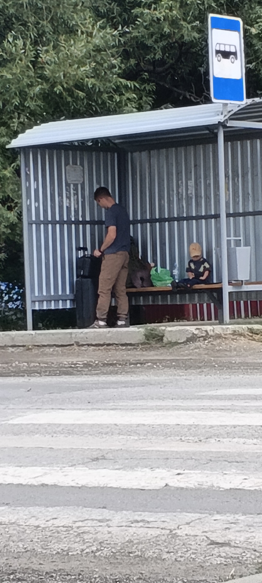 The image shows a bus stop in the city of Kargat, Russia. A man wearing a blue t-shirt and brown pants stands next to two black suitcases. He appears to be looking down at one of the suitcases, possibly adjusting something on it. To his right, a young child wearing a yellow cap and a patterned shirt sits on a wooden bench. The child is looking down at something in their hands. There is a green plastic bag on the bench next to the child. Behind the figures, the bus stop shelter has corrugated metal walls and a white roof. A blue and white bus stop sign is visible above the shelter on the right side of the image. The foreground shows a crosswalk and the rough ground of the street. Trees are visible in the background.
