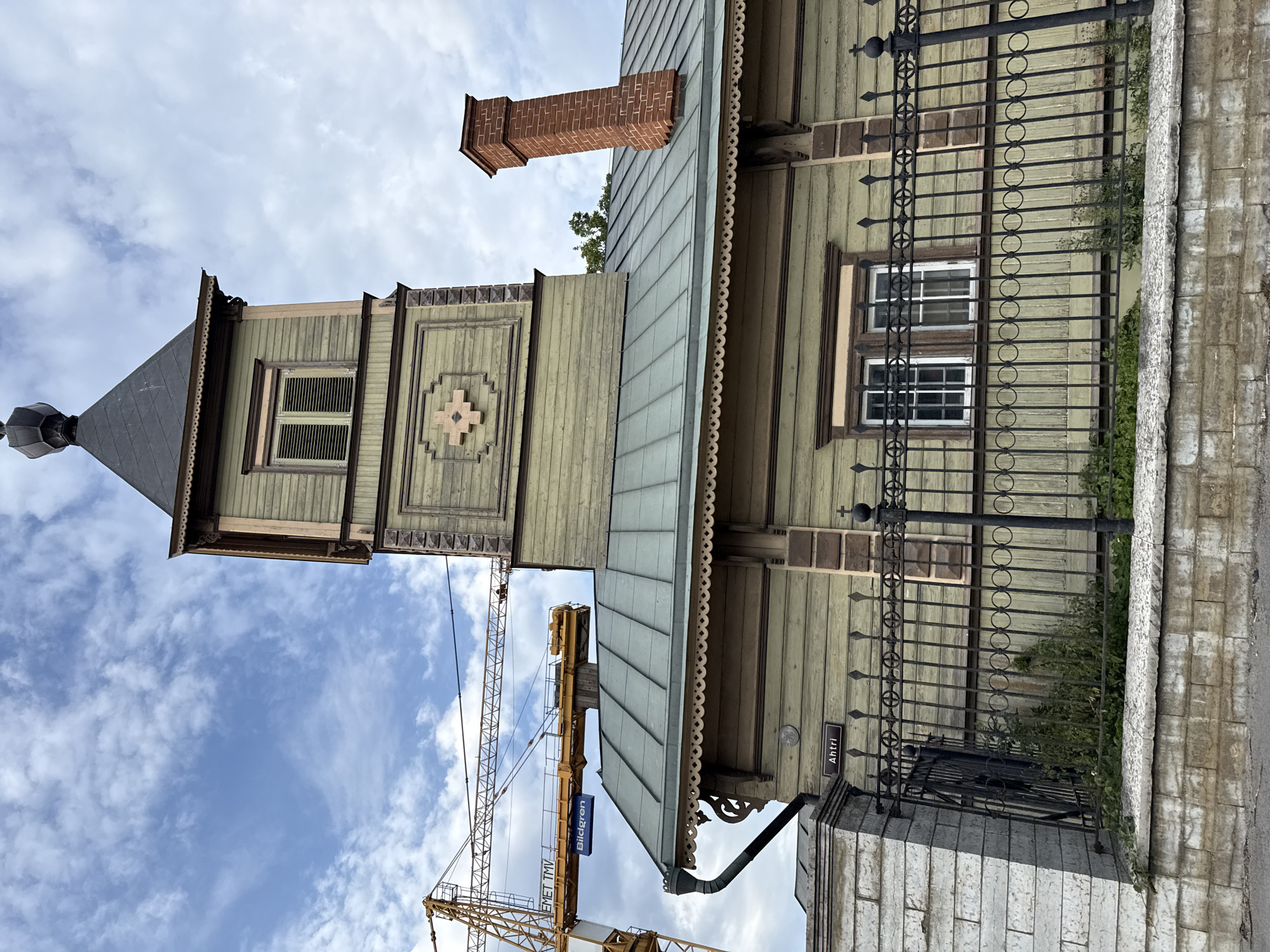 A low-angle outdoor shot captures a light green wooden building with a dark gray roof, possibly a church, under a partly cloudy sky. The building features a prominent bell tower with a dark gray, multi-faceted roof topped by a finial. The tower has a louvered window and decorative trim. A red brick chimney rises from the main roof. A black metal fence with decorative circles and pointed finials separates the building from a stone wall and sidewalk. In the background, a yellow construction crane with the text "Bildgren" is visible against the sky. A street sign reads "Ahtri." The overall scene suggests an urban or historical setting undergoing renovation or construction.