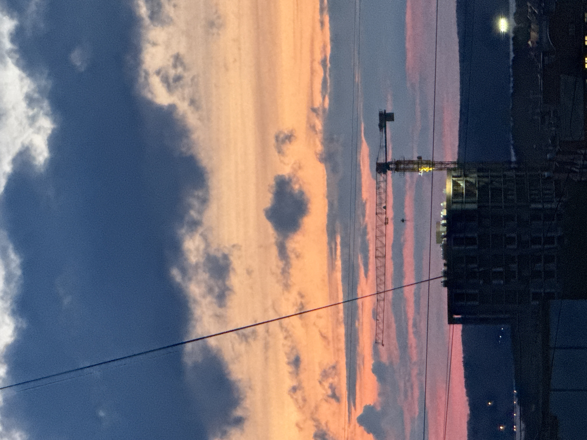 The image captures a vibrant sunset or twilight scene over Chaikovsky, Russia, characterized by dramatic cloud formations and urban infrastructure. The sky dominates the upper two-thirds of the frame, painted with striking hues of deep blue, soft orange, and pink, interspersed with large grey and white clouds. Patches of brighter, intense orange and pink suggest the sun's low position.
In the mid-ground and foreground, a prominent construction site is visible. A tall, skeletal tower crane stands illuminated with a yellow light, indicating ongoing work or safety lighting during dusk. Adjacent to the crane, the dark silhouette of a multi-story building under construction rises. Its frame and some windows are visible, with a few interior lights glowing, suggesting activity or occupation.
Several thin, dark lines, likely power lines or cables, stretch horizontally across the frame, adding an element of urban grit to the natural beauty of the sky. In the far distance, faint lights can be seen, hinting at other structures or parts of the city. The overall atmosphere is one of serene natural beauty juxtaposed with the ongoing development of an urban landscape. No people are visible, nor is there any discernible text.