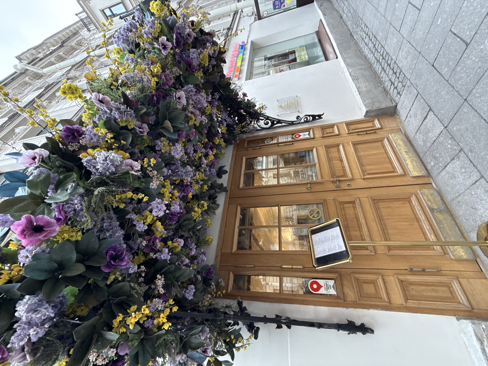 The image depicts the entrance to a building adorned with an abundant floral arrangement of artificial purple and yellow flowers cascading from above. The entrance features double wooden doors with glass panes and a signpost with a menu in front of them. A red marker with Russian text is visible on the door. To the right of the entrance, a store window displays colorful items and signage. The building appears to be in a city, with architectural details suggesting an older European style, and the pavement is paved with stone tiles. A street sign with Russian text, "Небываемо вкусно" (Unbelievably tasty), is visible above the store window. This is the entrance to the restaurant "Небываемо вкусно" on Bolshaya Morskaya Street in Saint Petersburg, Russia.