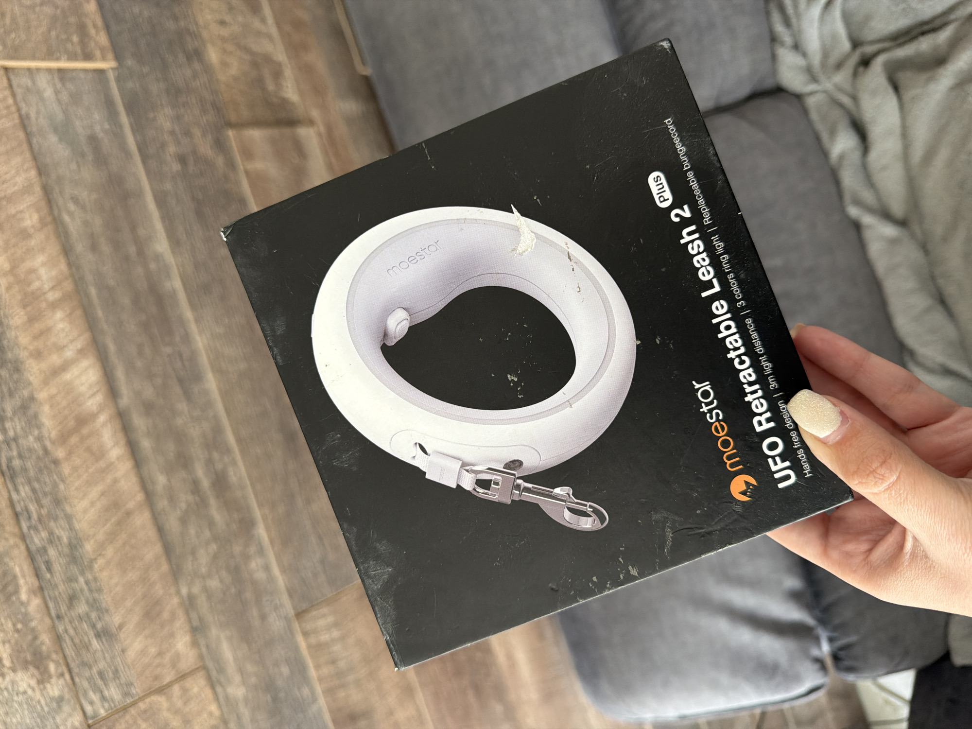 The image shows a box for a "moestor UFO Retractable Leash 2 Plus". The box is primarily black with an image of the white leash itself, which has a circular design. The text on the box indicates features such as "Hands free design", "3m light distance", "3 colors ring light", and "Replaceable bungeecord". A hand with a painted fingernail is visible, holding the edge of the box. The box is placed on what appears to be a wooden floor. In the background, part of a gray sofa is visible. There is no indication of the city of Burgas, Bulgaria in the image.
