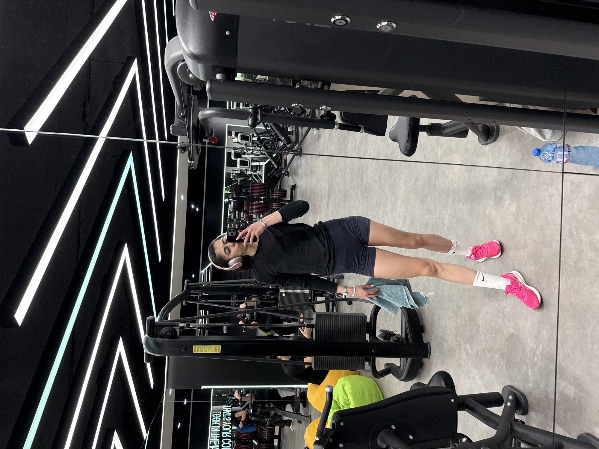 A young woman is taking a selfie in a mirrored wall at a gym in Sofia, Bulgaria. She is wearing a black top, dark shorts, white socks, and pink sneakers. She also has headphones around her neck and is holding a light blue towel. In the reflection, there is gym equipment, weights, and text. The wall behind the mirror has black and white geometric patterns illuminated by neon lights. A water bottle is visible near the top of the mirror.