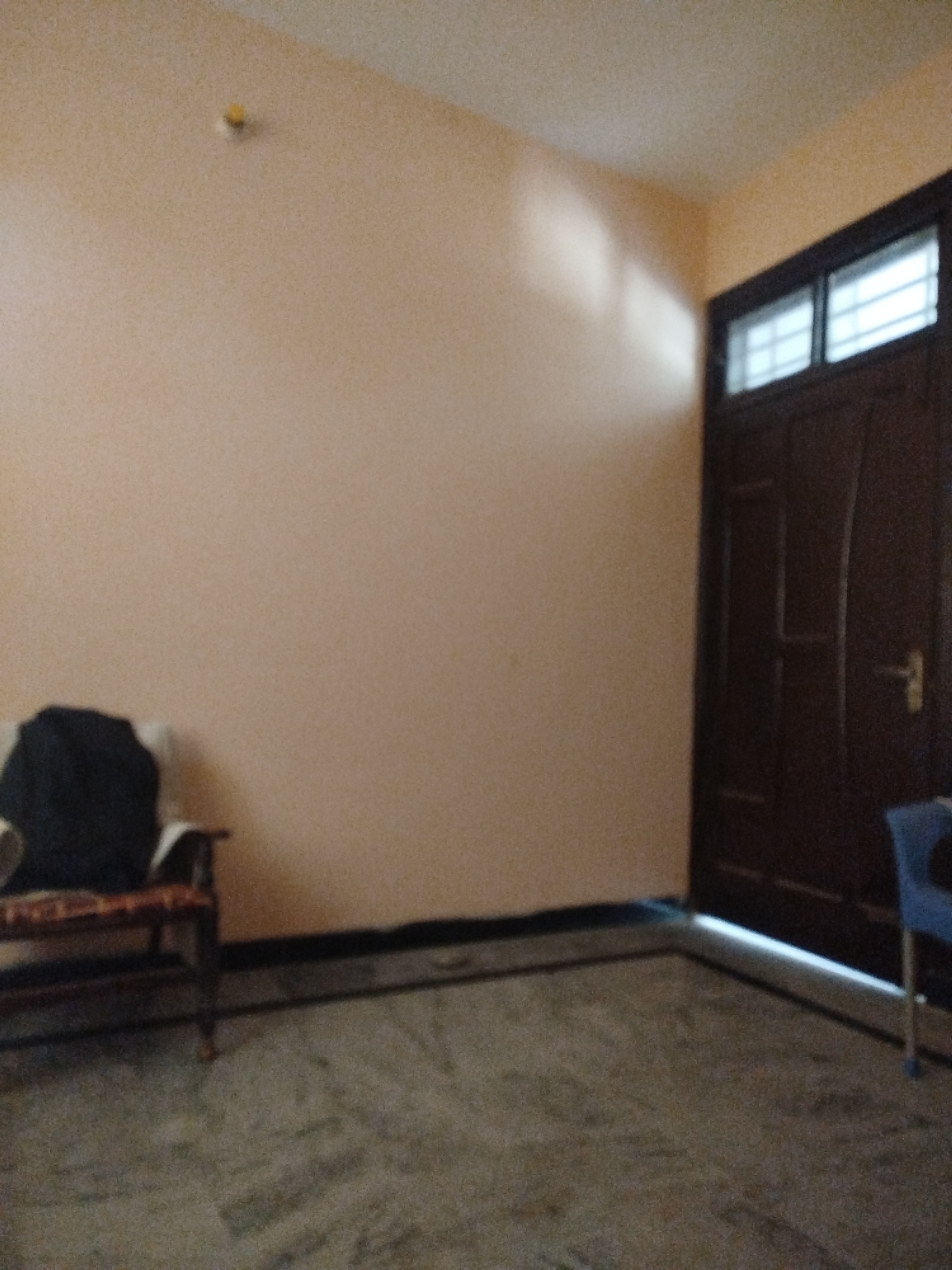 The image depicts an interior scene of a room with light peach-colored walls and a marble-patterned floor. On the left side of the frame, a dark-colored armchair with a cushion and a black bag is visible. A small yellow object is affixed to the wall near the ceiling. To the right, a large, dark wooden door with two rectangular window panes at the top is present. A blue chair is partially visible in the lower right corner. The scene is likely a room in a residential or commercial building in Lassan, Pakistan.