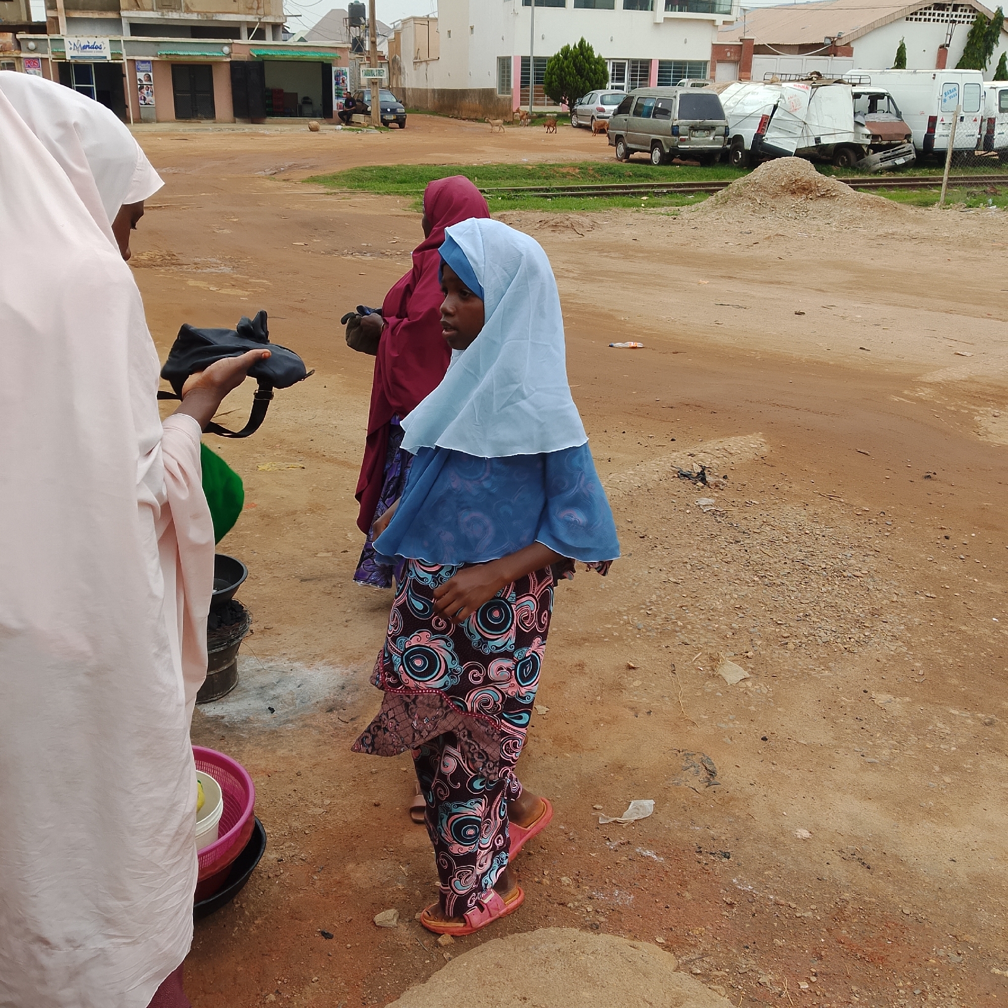 In a dusty, outdoor urban environment, three individuals are visible. The most prominent subject, a young female wearing a light blue hijab and a patterned blue and pink dress, stands facing the right side of the frame. To her left, another female is mostly obscured by the foreground subject, wearing a dark red hijab and a dark patterned skirt. Partially visible on the far left is a third individual, appearing to be an adult, draped in a pale pink garment, holding a dark bag. The ground is primarily composed of reddish-brown dirt and gravel. In the background, a damaged white van is parked amongst other vehicles, suggesting a roadside or an area with parked vehicles. The scene appears to be in a commercial or residential area, with buildings visible behind the subjects.