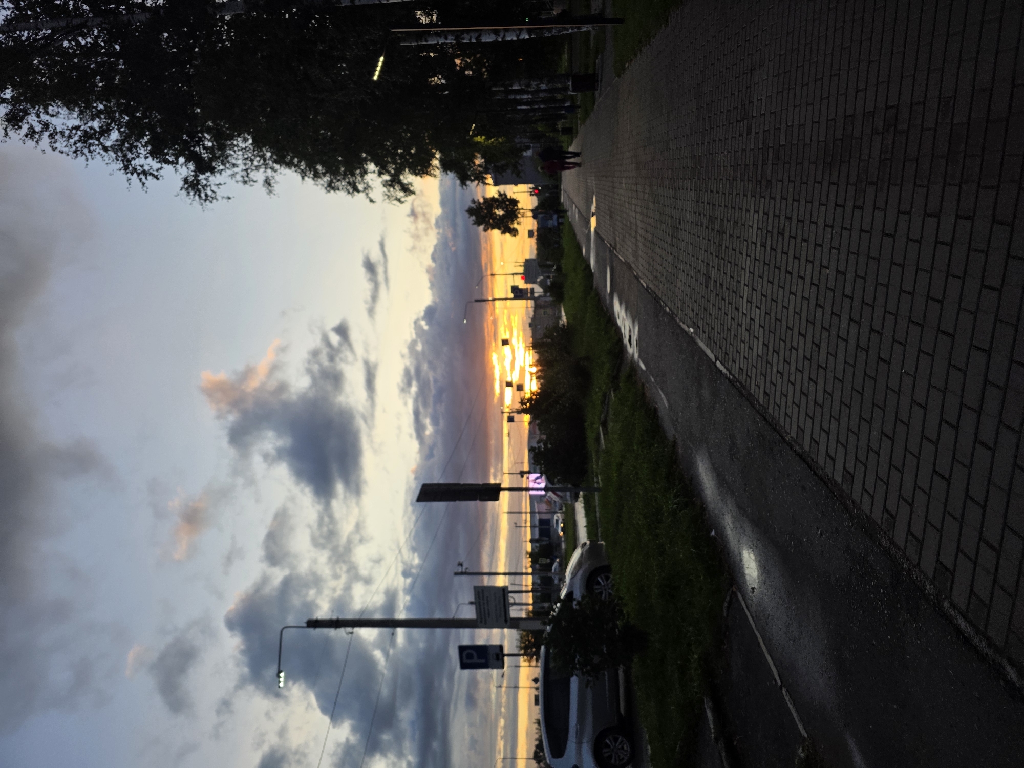 The image captures an urban scene at dusk, likely in Chaikovsky, Russia, given the presence of birches and the overall landscape. A paved walkway, made of dark rectangular bricks, stretches into the distance, bordered by a grassy verge on the left and a line of trees on the right. The sky is a blend of twilight hues, with soft clouds tinged with pink and orange from the setting sun, which is partially obscured by buildings and trees on the horizon. A streetlamp with three lights stands on the left side of the walkway, illuminating the darkening sky. Adjacent to the walkway is a wet asphalt road where a silver car is parked, and further down, a few more vehicles are visible, their headlights on. Two figures, silhouetted against the fading light, are walking away from the viewer on the walkway. The environment suggests a tranquil evening, with remnants of recent rain evident on the road and a puddle reflecting the sky.