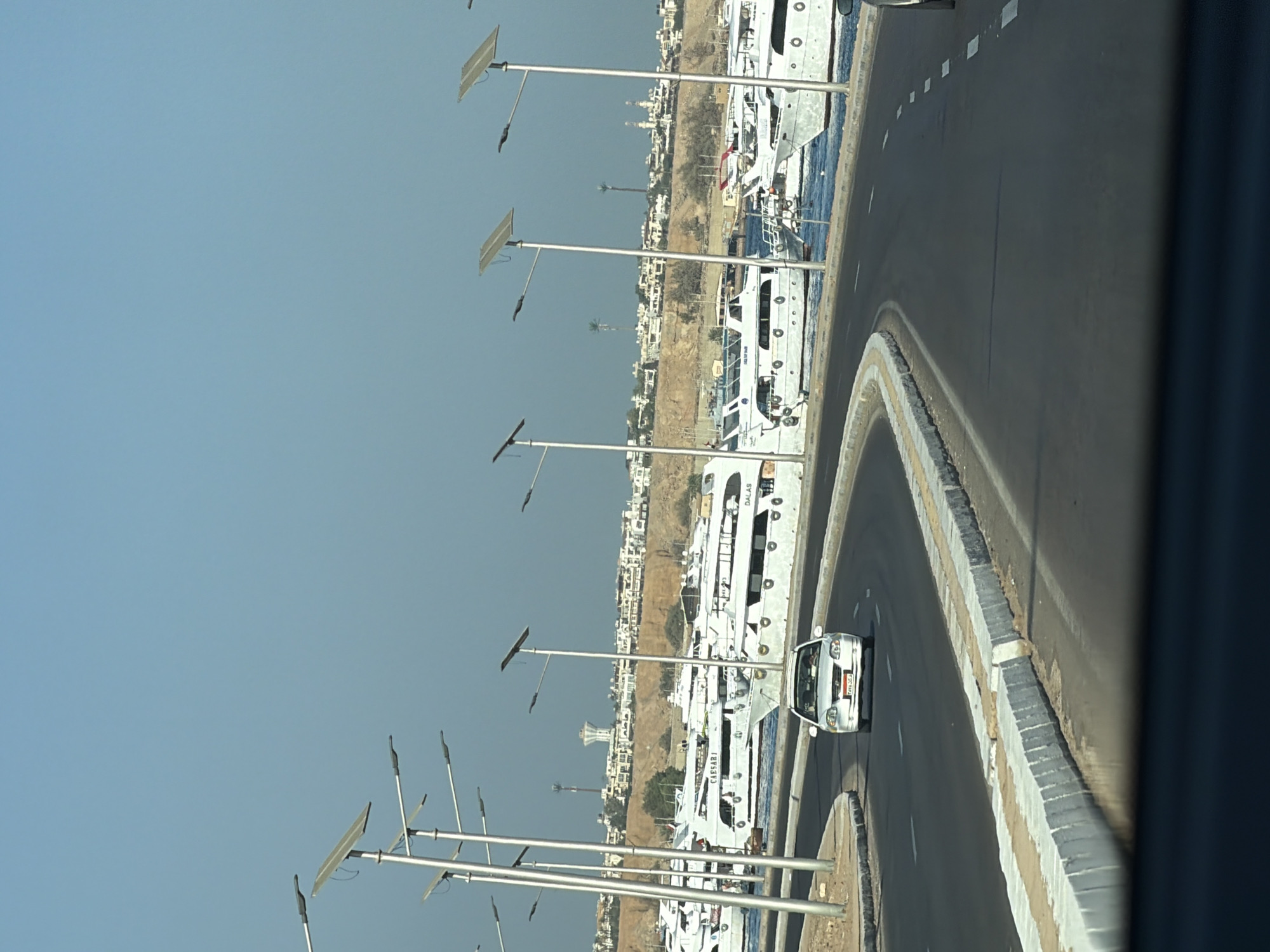 The image depicts a view of a coastal road in Sharm El Sheikh, Egypt. A white sedan is driving on a curved asphalt road with white lane markings. On the left side of the road, there is a concrete barrier with a light-colored curb. Beyond the barrier, a marina is visible, filled with numerous white yachts docked in the water. In the background, a town is built on a hillside, characterized by white buildings and some vegetation. Several solar-powered streetlights line the road, equipped with large panels. The sky is clear and blue, indicating fair weather. The overall scene captures a typical day at a harbor city.