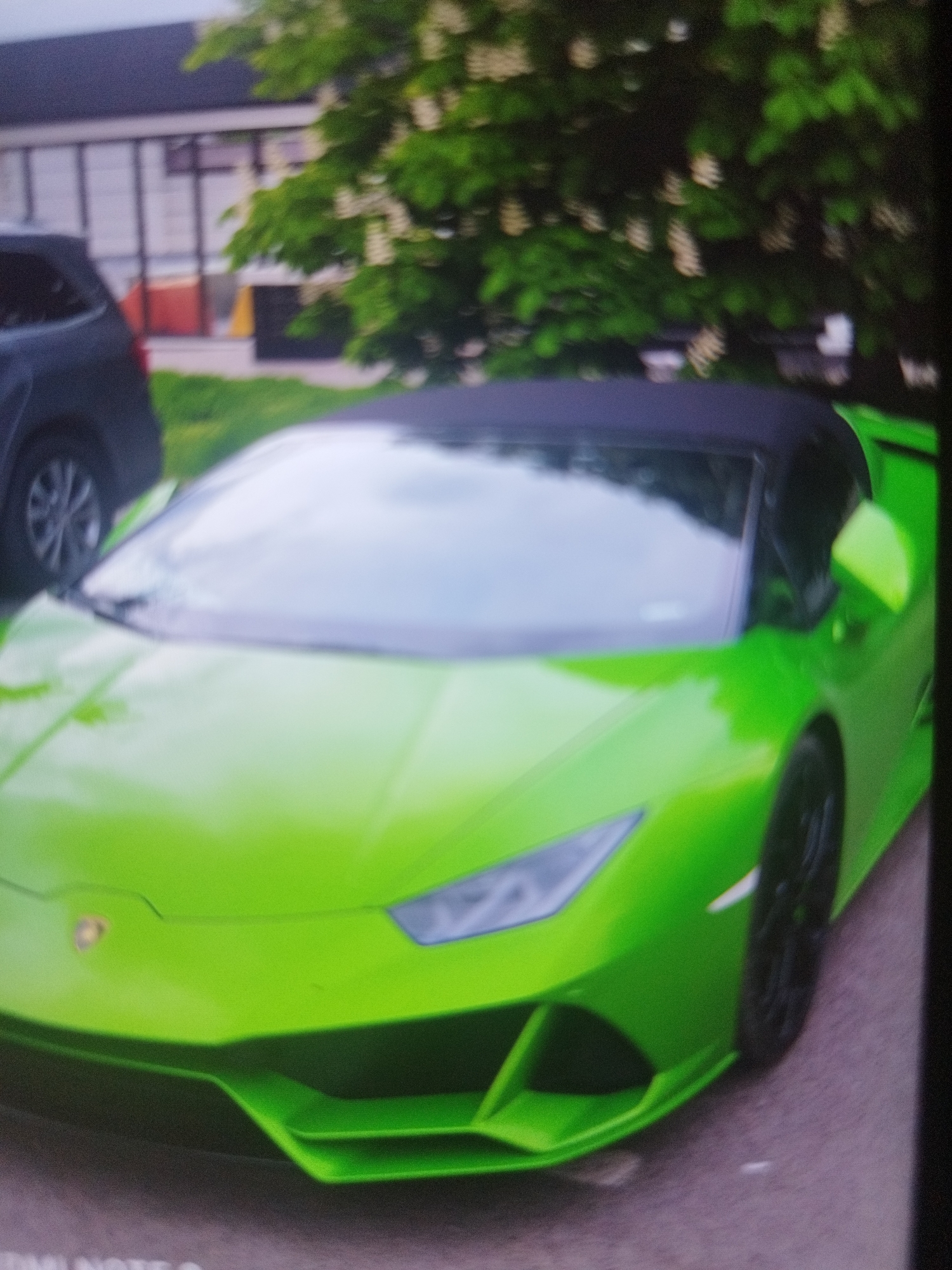 A slightly blurry, low-resolution image shows a bright green Lamborghini Huracan Spyder parked on a street. The car has a black soft top. The front of the car is clearly visible, showing the headlights and front bumper. Part of a dark SUV is visible to the left of the Lamborghini. Behind the car, there is some greenery, and a building with a distinctive multi-colored facade can be seen in the background.
While the specific location can't be determined with certainty from the provided photo alone, the building's architecture and color scheme could potentially be used to identify the area within Plovdiv, Bulgaria.