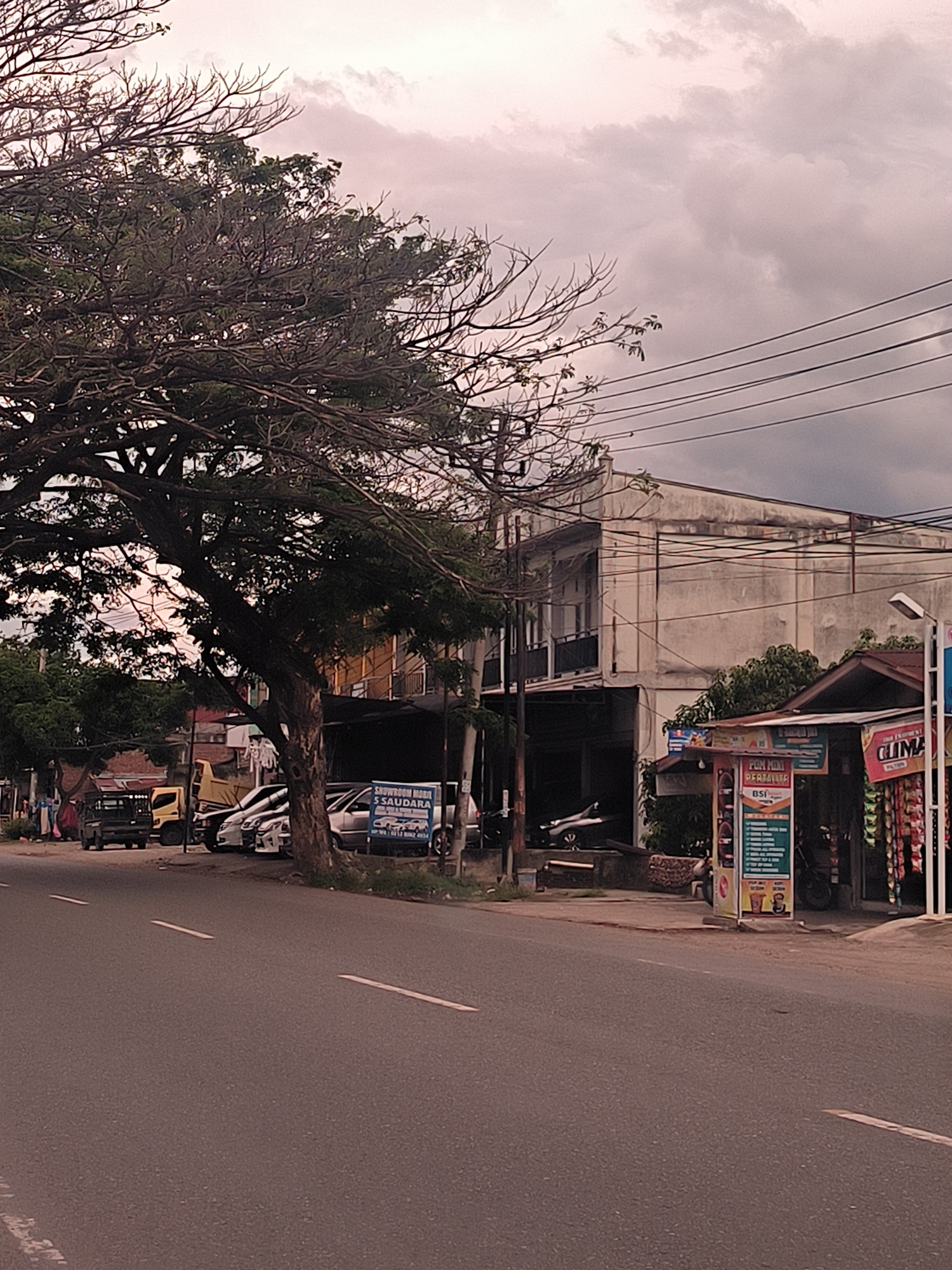 The image depicts a quiet street scene in Ujong Duabelas, Indonesia, under an overcast sky, suggesting either a very cloudy day or late afternoon. A large, sprawling tree with both leafy and bare branches dominates the left side, casting shade over the road and parked vehicles.
The foreground features an asphalt road with white dashed lane markings. Along the left side of the road, several vehicles are parked, including a dark pickup truck, a yellow dump truck, and multiple sedans, some appearing to be new or used cars. A blue sign reads "SHOWROOM MOBIL 5 SAUDARA" (Car Showroom 5 Brothers/Siblings), with a car logo and contact details, confirming the presence of a car dealership or used car lot.
On the right side, the street is lined with commercial buildings and smaller stalls. A prominent two-story building with a weathered, light-colored facade and a second-floor balcony stands further back. In front of it, closer to the road, are smaller, single-story structures. Numerous power lines crisscross the upper right portion of the image, connected to utility poles.
A vertical banner advertisement is visible near one of the stalls, displaying "PUM MINI PERTALITE" (Mini Pertalite Fuel) and "BSI," along with a list of services or products on a green background. Above it, a smaller red sign reads "ES KRIM" (Ice Cream). Further to the right, a red banner partially visible says "G_LIMA," likely an advertisement for a shop selling snacks or goods, which are displayed beneath it. One person appears to be seated near the "PUM MINI PERTALITE" stall.
The overall mood is calm and subdued, with no apparent hurried activity.