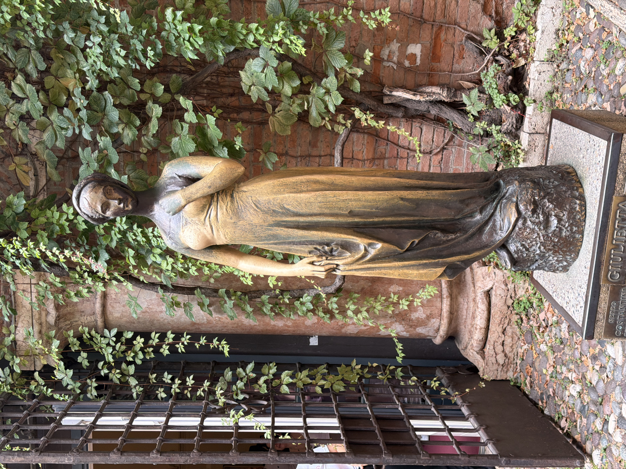 The image shows a bronze statue of Juliet, a character from Shakespeare's play Romeo and Juliet, in Verona, Italy. The statue is located in Juliet's House (Casa di Giulietta), a medieval building in Verona, believed to be the home of Juliet. The statue stands on a pedestal in the courtyard, surrounded by ivy-covered brick walls. A large metal gate and a column are visible to the left of the statue. The ground is paved with cobblestones and scattered with fallen leaves. The statue is depicted in a flowing gown, with one hand on her chest and the other holding her dress.
