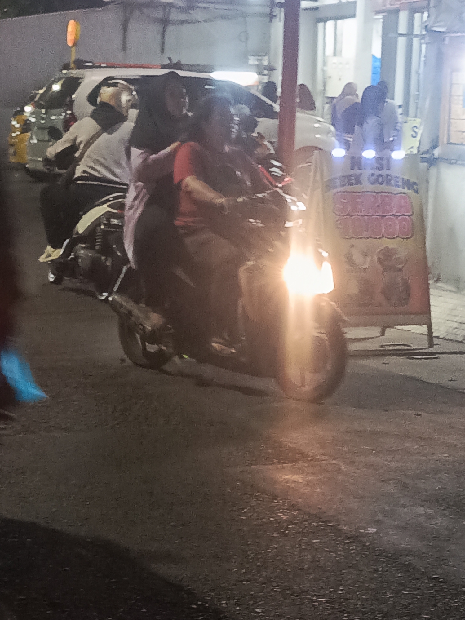 In Surabaya, Indonesia, at night, a motorcycle carrying three people, two adults and a child, is visible from the side. The adult in the front is wearing a red shirt, the adult behind is wearing a dark hijab and grey jacket, and the child is on the far back. The motorcycle's headlight is on and illuminated. Behind the motorcycle, a sign is visible, with the text "NASI BEBEK GORENG" and "SAMBA BALADO" written on it, indicating it's a food stall. A white car with a yellow stripe is also parked to the left of the motorcycle. The environment appears to be a street at night with artificial lighting.