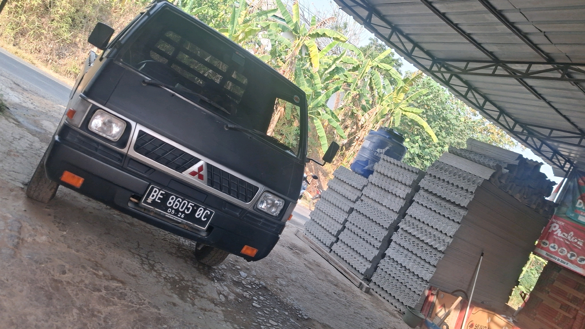 The image captures a dark-colored truck parked outdoors, tilted on a dirt or gravel surface. The truck is positioned under a metal-roofed structure, and to its right, a large stack of corrugated roofing materials is visible. Behind the truck and to the left, lush green foliage and trees suggest a rural or semi-rural environment. A road is partially visible to the left of the truck. The license plate on the truck reads "BE 8605 OC". The scene appears to be in a storage or roadside area, possibly a business related to construction materials. The lighting indicates it is daytime, with sunlight casting shadows. The specific location is identified as Karang Sari, Indonesia.