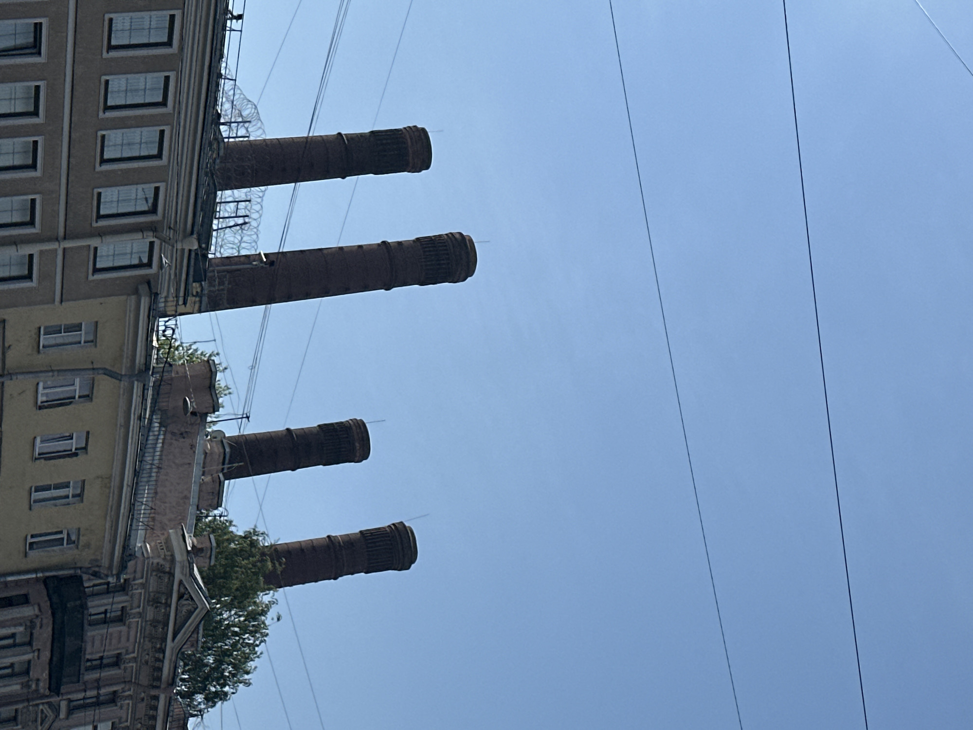 The image depicts a building in Saint Petersburg, Russia, with four prominent chimneys extending upwards. The building's facade is beige and features rows of windows. Power lines stretch across the clear blue sky in the background. Some greenery is visible around the base of the chimneys and on the building's structure. The scene is oriented with the sky occupying a significant portion of the frame.