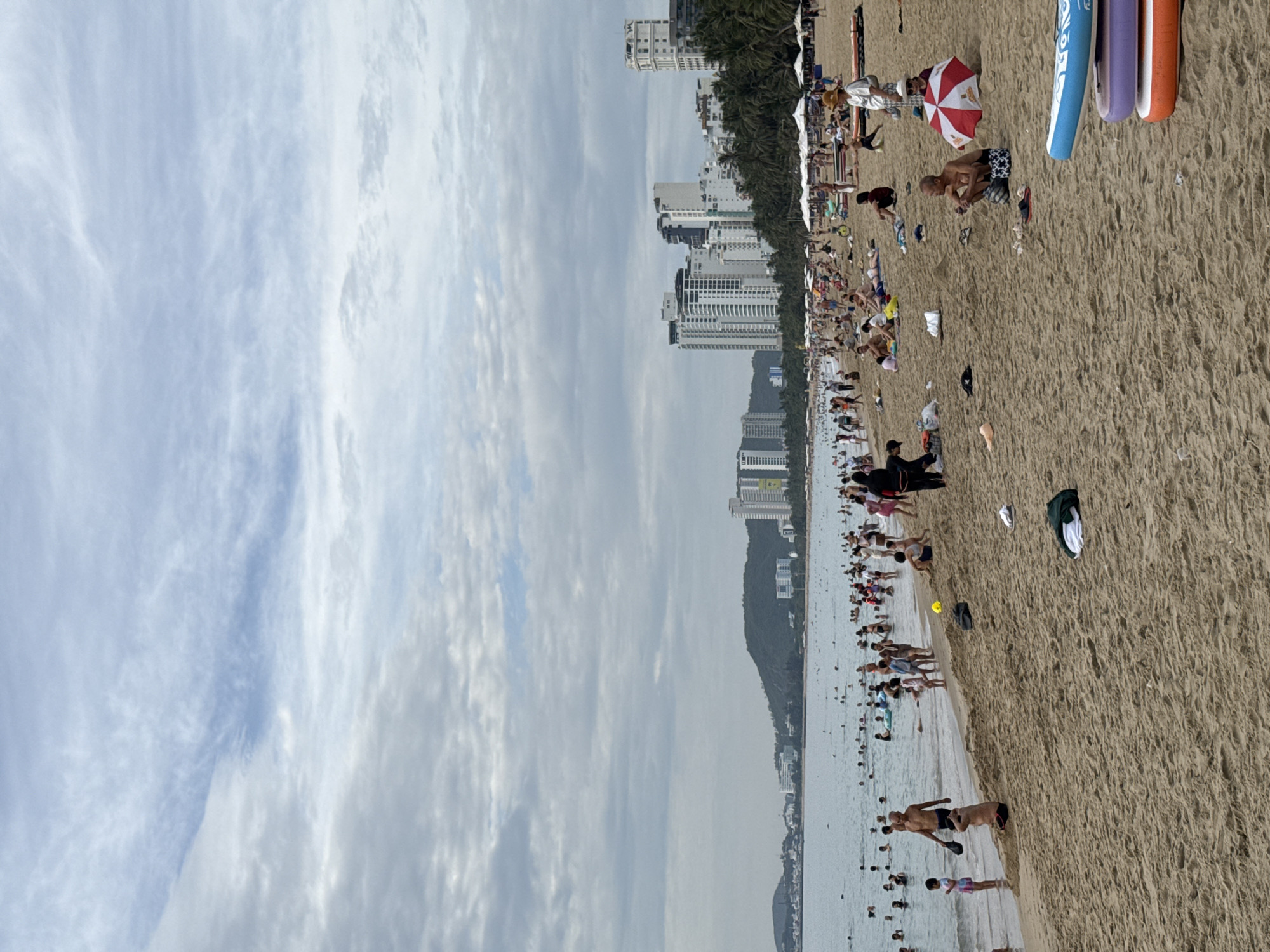 The image depicts a crowded beach in Nha Trang, Vietnam, on a cloudy day. The foreground is dominated by a sandy beach with numerous people sunbathing, relaxing, and walking. Several beach chairs and umbrellas are set up, providing shade. In the middle ground, a large body of water is filled with swimmers and people wading. The background features a coastline with palm trees and a dense collection of high-rise buildings, characteristic of a coastal city. The sky is overcast with scattered clouds. Several paddleboards are stacked on the beach in the lower right corner.