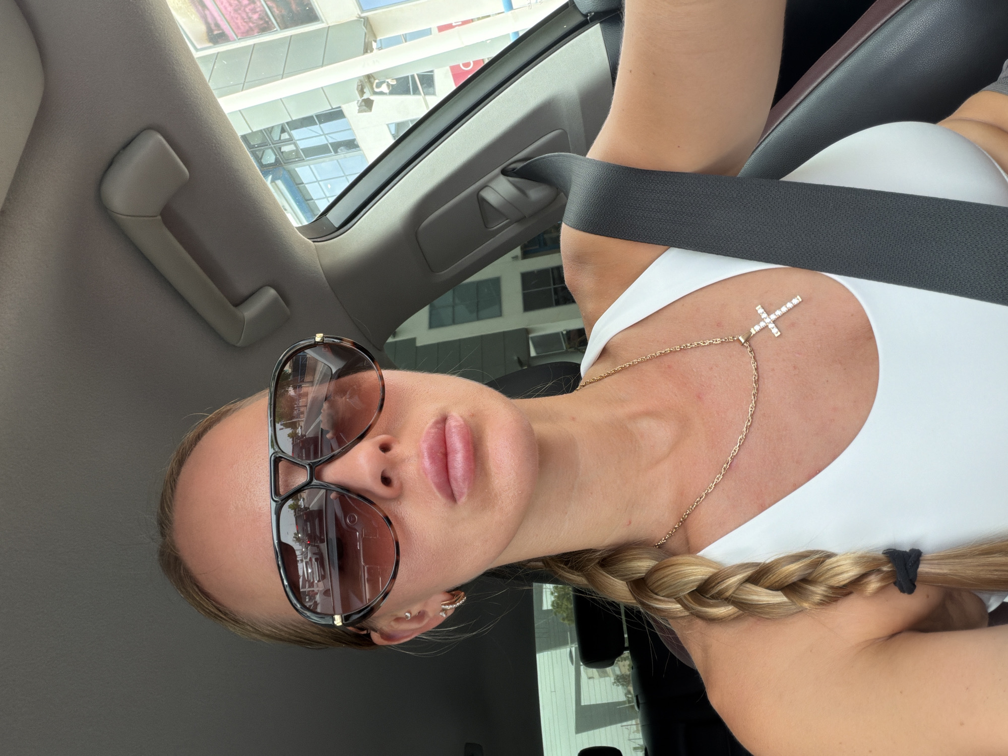 A blonde woman with her hair in a braid is driving a car in Finestrat, Spain. She is wearing sunglasses, a white tank top, and a gold necklace with a cross pendant. The car's interior is visible, including the seatbelt and a grab handle. Outside the car, buildings and other vehicles can be seen.