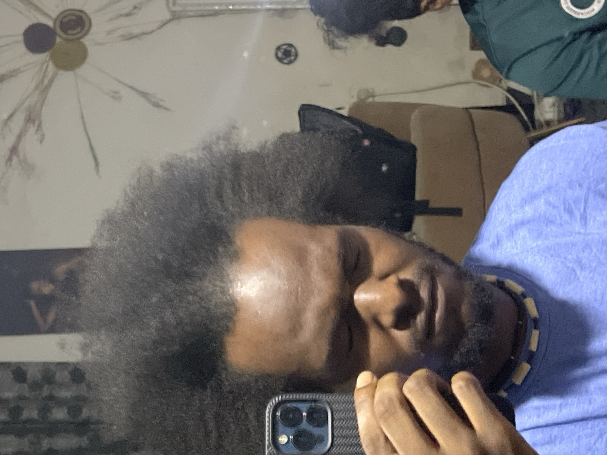 A medium-shot, slightly rotated, indoor image shows a dark-skinned person with an afro hairstyle and a blue t-shirt. The person is holding a smartphone with a case that has three circular lenses in the lower portion of the frame. The person is wearing a beaded necklace. Background objects include a tan chair, a black bag, and what appears to be wall decorations. The location in Kuchingoro, Nigeria is not discernible from the image.
