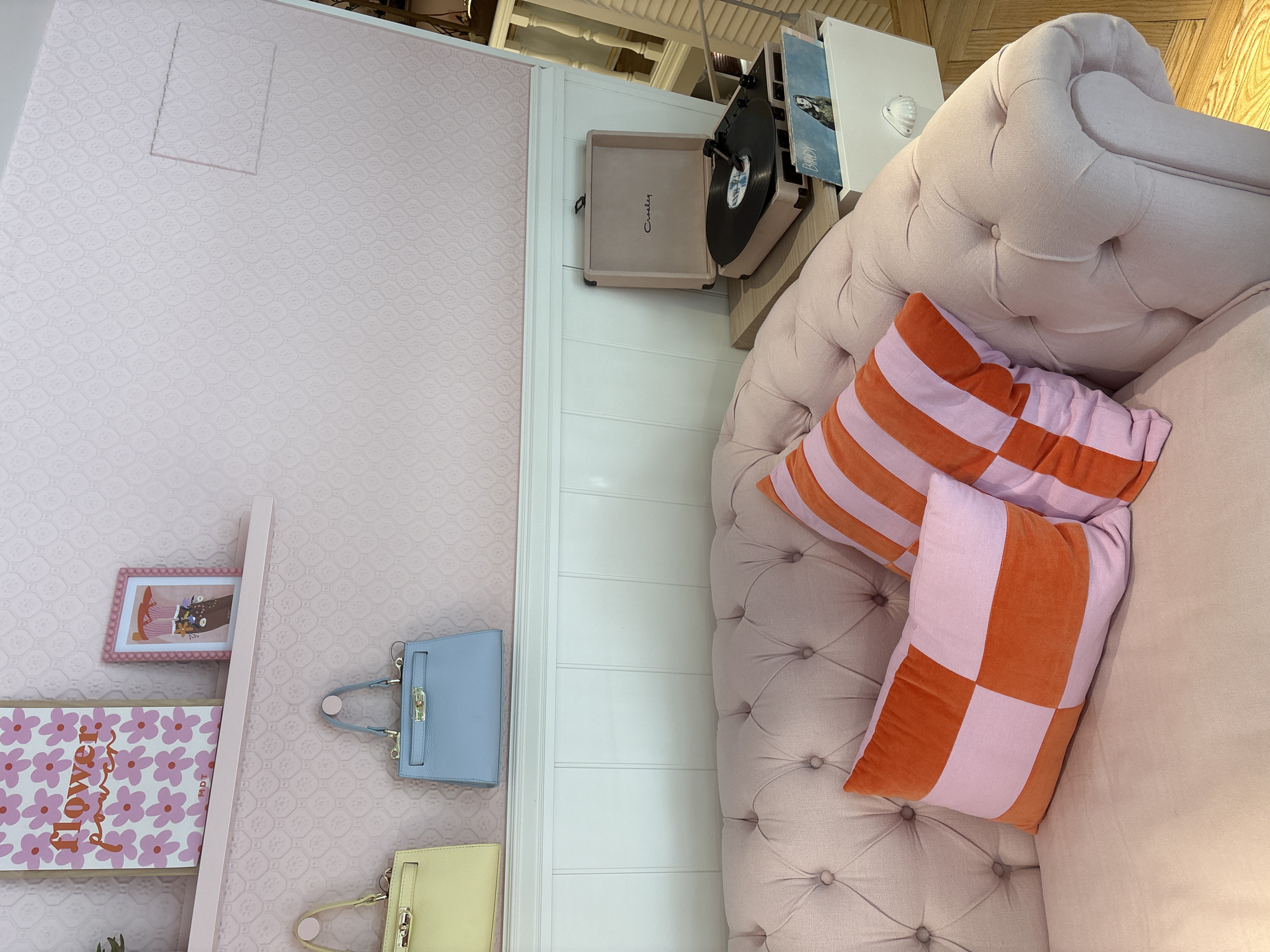 This is a close-up, eye-level shot of a pink tufted couch with two decorative pillows. The pillow on the left is a checkerboard pattern of pink and orange squares. The pillow on the right has horizontal pink and orange stripes. Behind the couch, a pale pink wall with a subtle floral pattern is visible. A white wooden wainscoting runs along the bottom of the wall. On the wainscoting, to the right, sits a pink record player with a record on it. To the right of the record player, a white drawer with a shell-shaped handle is slightly pulled out, revealing a record with the name "BIRDY" printed on it. On a shelf above the wainscoting, two handbags are hanging: one is pale yellow, and the other is a pale blue. Above the handbags, a framed picture with pink flowers and the words "flower power" is displayed, along with another smaller framed picture. The floor is made of light-colored wood planks. The scene appears to be an indoor setting, possibly a living room or a styled retail space, and there is no indication of it being in Sanxenxo, Spain.