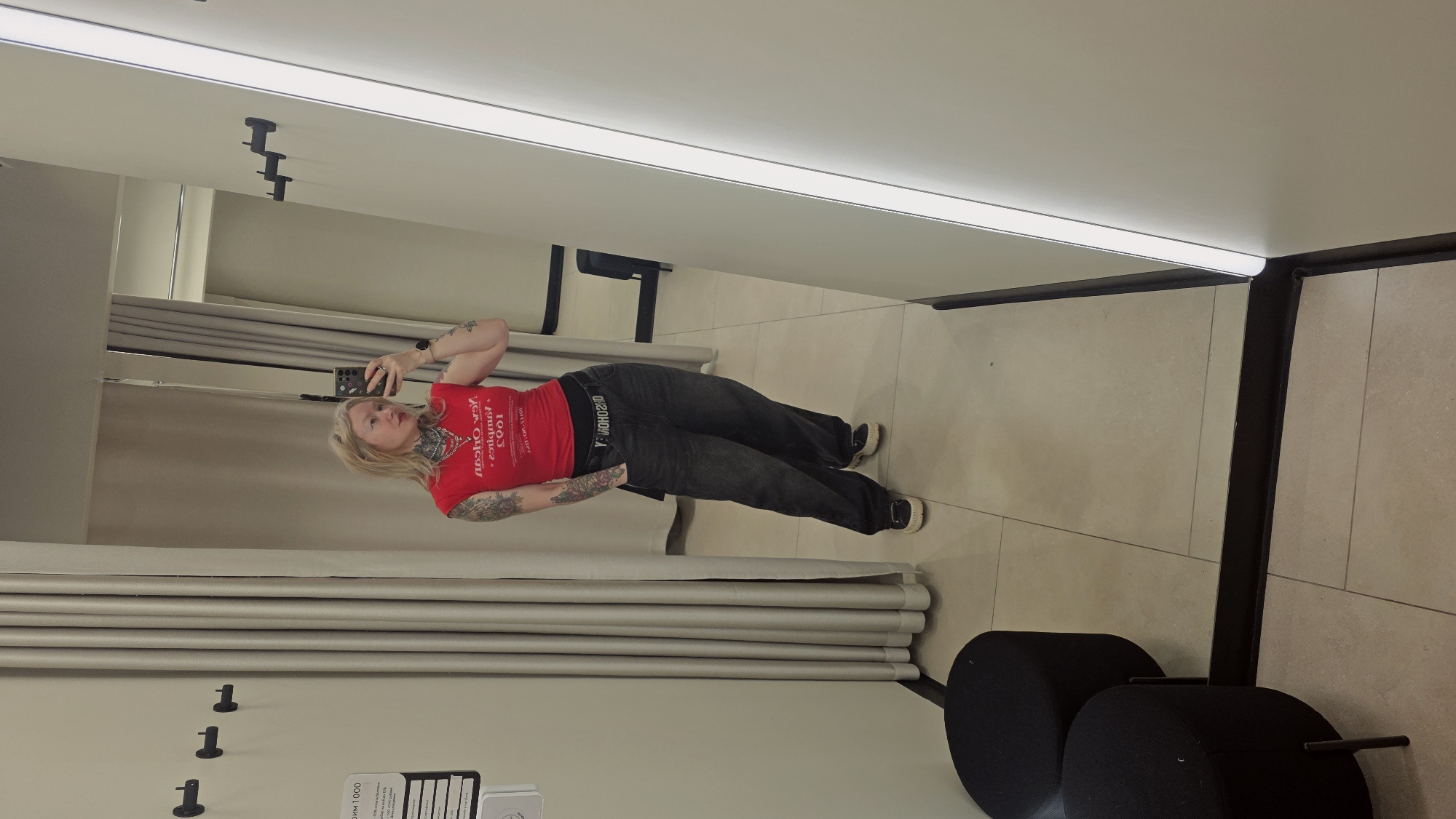 A blonde woman, appearing to be in her 20s or 30s, is captured taking a mirror selfie in what appears to be a fitting room or clothing store in Saint Petersburg, Russia. She stands centered in the frame, holding a dark-colored smartphone with her right hand to photograph her reflection.
The woman is wearing a fitted, short-sleeved red top with white reversed text, and dark, loose-fitting jeans. She has a visible tattoo on her neck/chest area and extensive tattoos covering both arms. Her sneakers are black with white soles.
The setting is an indoor space, likely a retail environment, characterized by a large, full-length mirror that fills much of the frame. To the left, three black clothing hooks are mounted on a light-colored wall. Behind the woman, light-colored drapes or blinds are visible in the reflection. In the foreground to the right, two black, round, upholstered stools are present. The floor is tiled with light, rectangular tiles, and the ceiling features a long, linear light fixture.
Visible text includes the reversed "1003" and other smaller, unreadable text on her red top, and "YOHOOD" visible on the waistband of her pants. A partially obscured white information card on the wall to the left shows "1000" and some Russian text, further supporting the Saint Petersburg location context. The lighting suggests artificial indoor conditions. The woman's expression is neutral as she focuses on her phone.
