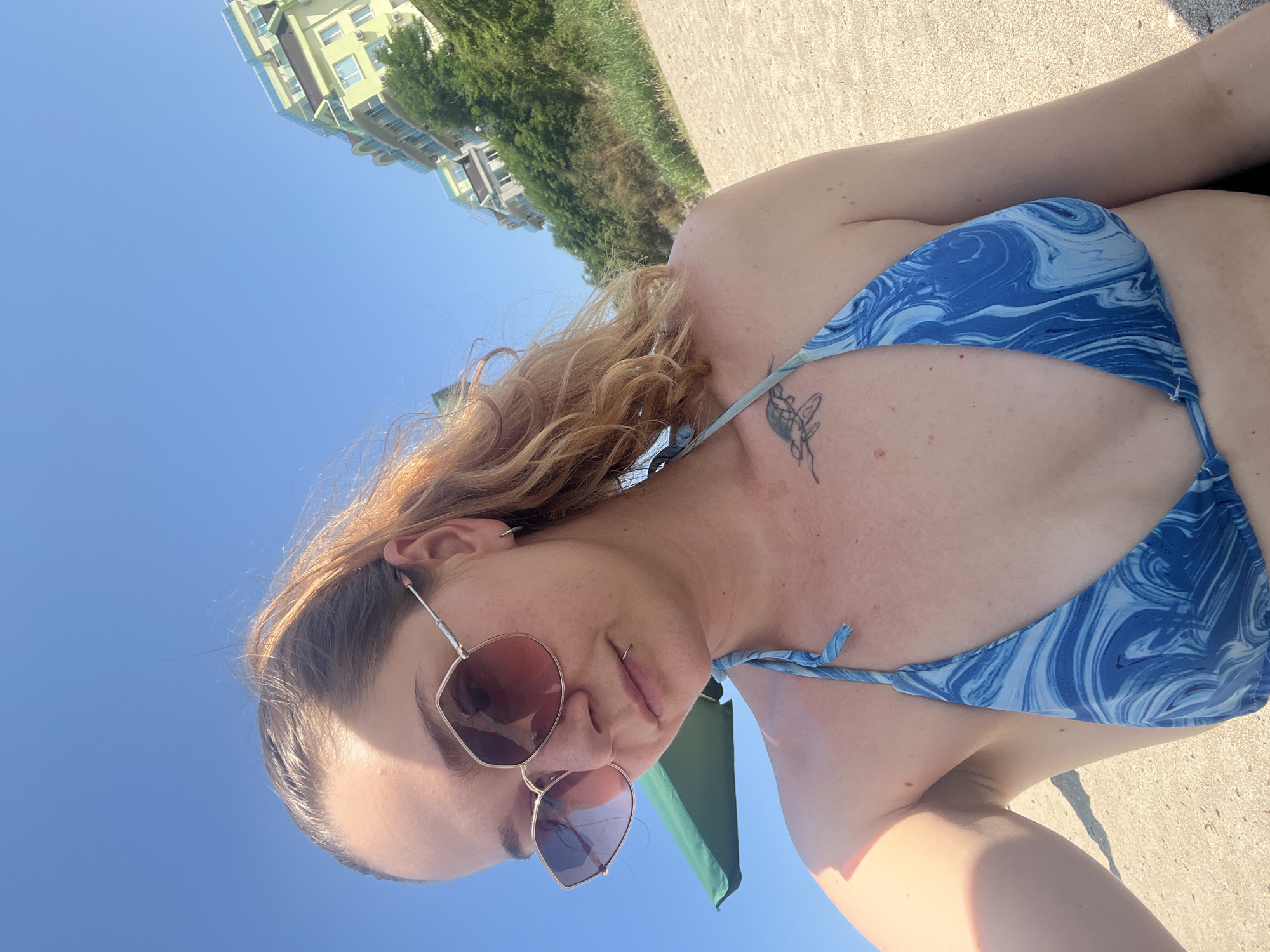 A selfie taken by a woman on a sandy beach in Lozenets, Bulgaria. She is wearing a blue and white marbled bikini top and large, pink-tinted sunglasses. Her hair is pulled back in a ponytail and is a reddish-blonde color. She has a piercing on her lower lip and a tattoo of a sea turtle on her chest. In the background, the clear blue sky is visible, along with a lush green hillside topped with a light yellow building. A portion of a green beach umbrella is also visible on the left side of the image.