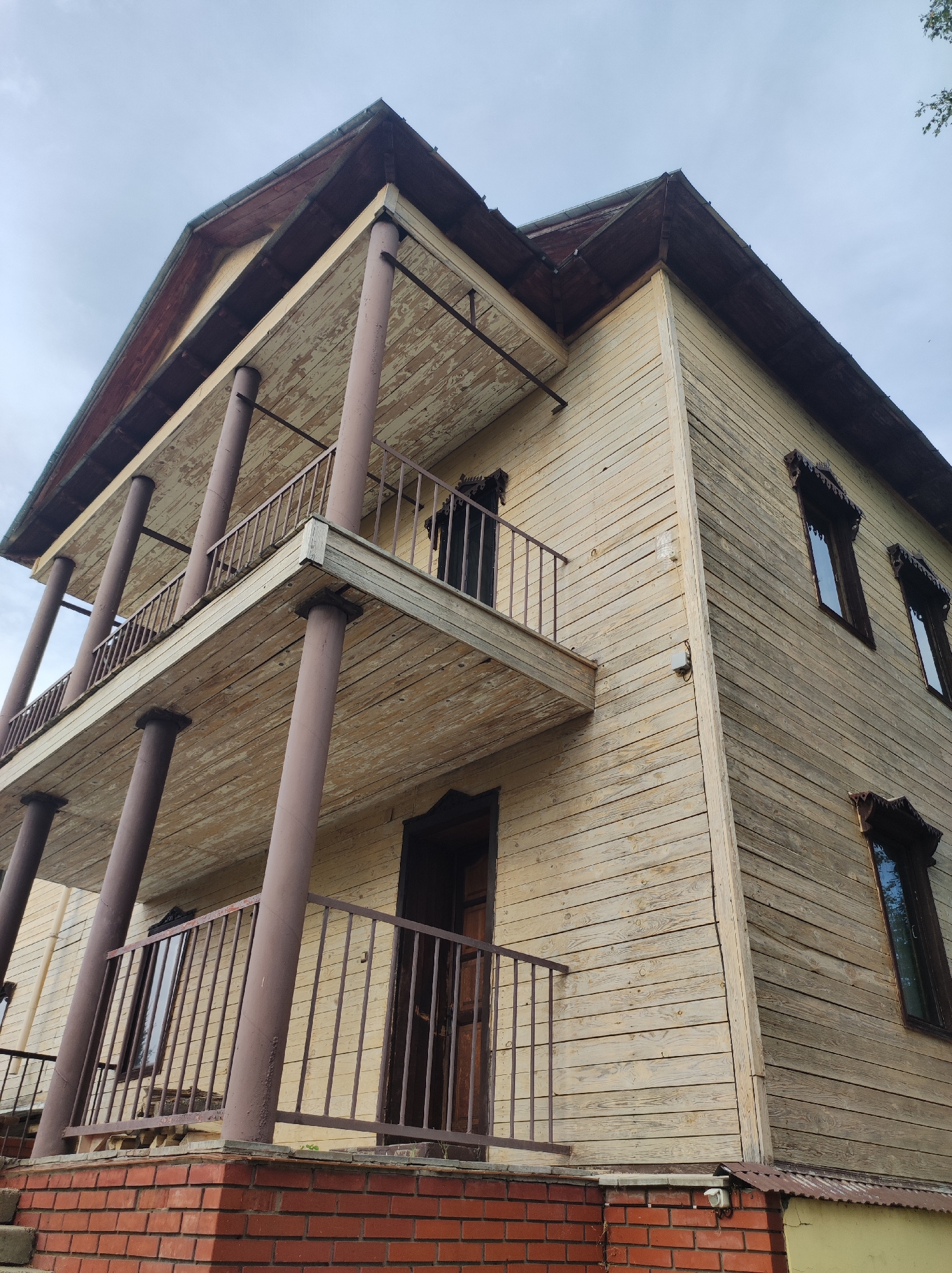 An exterior, low-angle, wide shot captures a weathered, light-colored wooden two-story building with a dark brown roof. The building features two balconies, supported by thick, cylindrical columns painted a reddish-brown. The balconies have metal railings. The wooden siding is horizontal and shows signs of peeling paint. Several dark-framed windows with decorative wooden trim are visible on the upper floor. The building rests on a foundation of red brick. The sky above is overcast with grey clouds. The scene is devoid of people or any visible activity. The image is likely from Zavod Mikhailovskii, Russia, given the architectural style suggestive of older rural or semi-rural Russian settlements.
