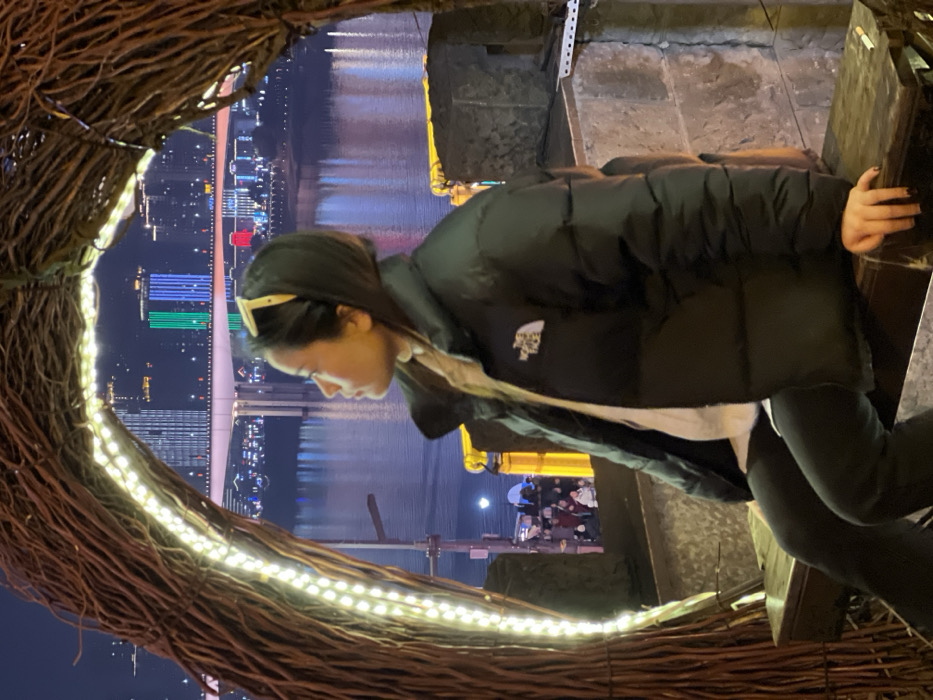 The image depicts a nighttime urban scene in Yuzhong District, Chongqing, China, overlooking a river and cityscape. In the immediate foreground, a young East Asian female is seated in profile, facing left, within a large, arch-shaped structure constructed from dark, intertwined branches or twigs. The inner curve of this structure is illuminated by a strip of white LED lights.
The female subject wears a black puffer jacket with a white "The North Face" logo on the left chest, over a light-colored top. She also has dark trousers and a light-colored headband or sunglasses pushed up onto her dark hair. Her left hand rests on the dark wooden surface of the bench she occupies. A cigarette butt is visible on this wooden surface next to her hand. She is gazing upwards and to the left, towards the distant cityscape.
In the middle ground, a wide river, likely the Yangtze River, reflects the numerous city lights. The illuminated Dongshuimen Bridge spans the river. Beyond the bridge, a dense urban skyline is visible, characterized by multiple high-rise buildings, many of which are lit, including some displaying prominent red, green, and blue rectangular lighting patterns. Below the bridge level, to the left of the main subject, a blurred group of people is visible in a public area. The overall scene is brightly lit by artificial sources against a dark night sky.