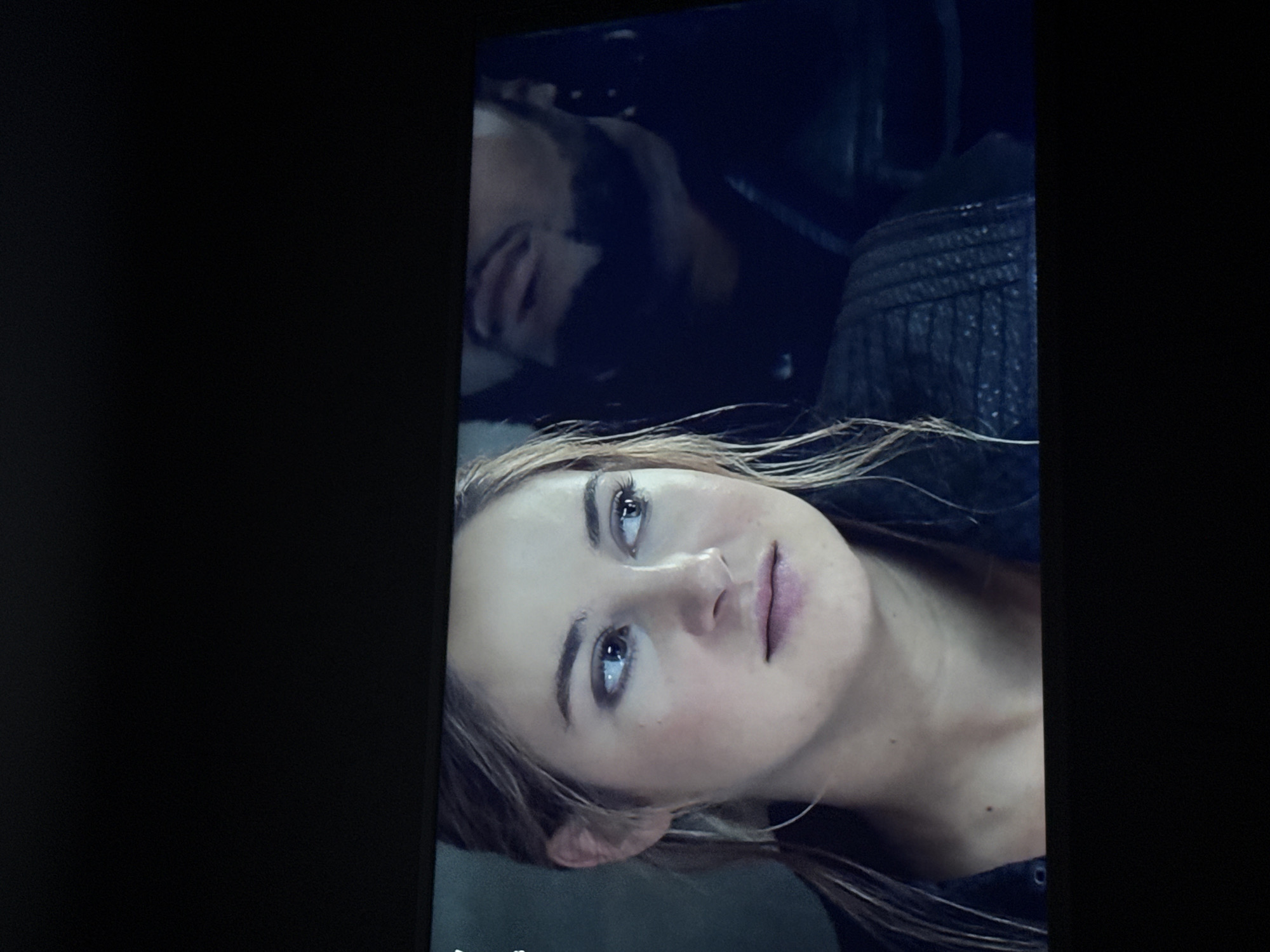 A medium shot of a television screen with a movie paused. The scene shows a fair-skinned woman with light brown hair, blue eyes, and wearing a black leather jacket. Above her is a blurry image of a bearded man, also wearing a black leather jacket. The background is dark, and the location is not identifiable. There is no evidence the image was taken in Mytishchi, Russia.
