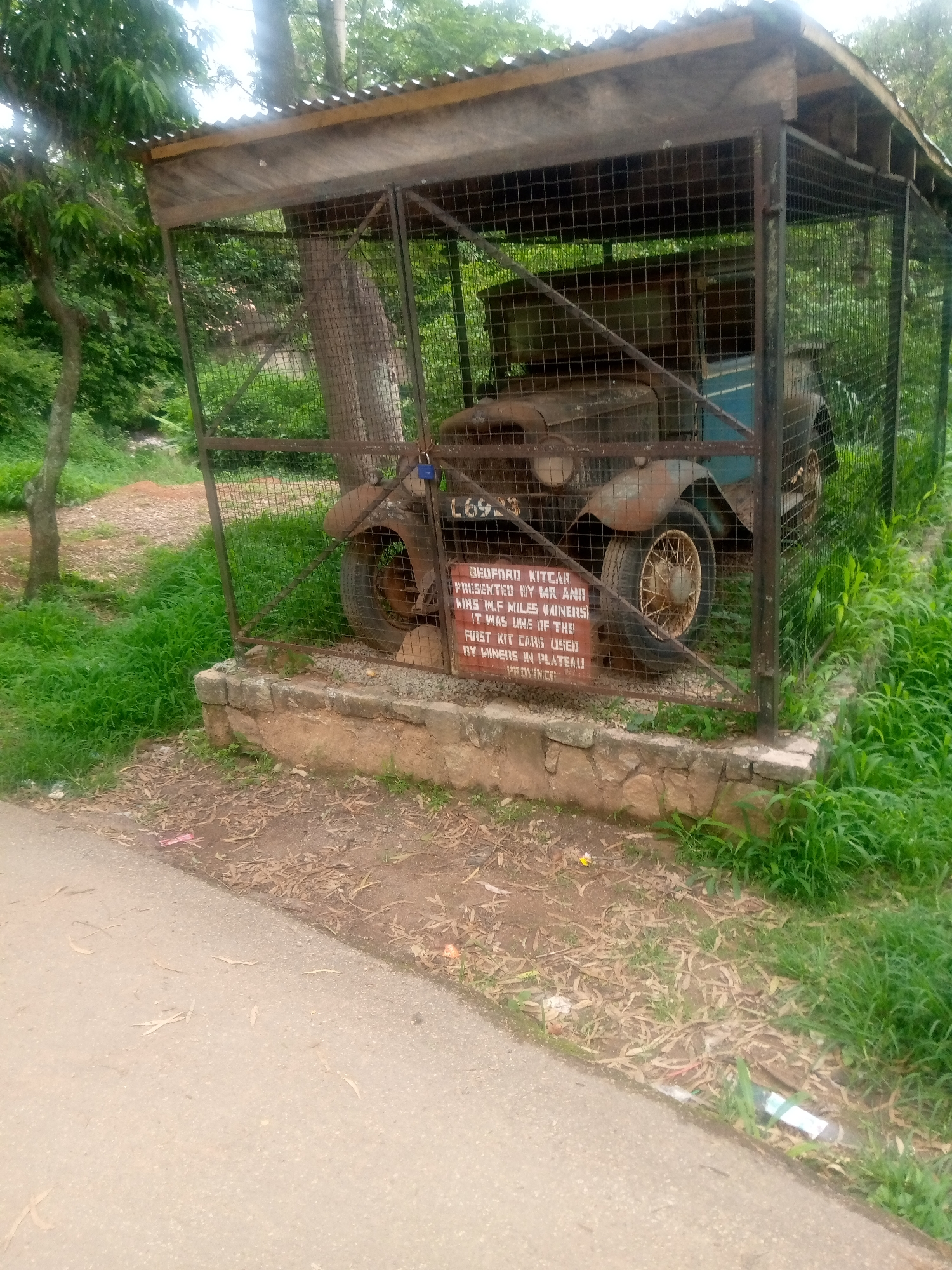 An old, rusty Bedford kitcar is housed in a protective cage with a corrugated metal roof. The car, featuring a blue interior and a license plate that reads "L6923," is in disrepair. A sign attached to the cage provides information about the vehicle: "BEDFORD KITCAR PRESENTED BY MR AND MRS H.F. MILES (MINERS). IT WAS ONE OF THE FIRST KIT CARS USED BY MINERS IN PLATEAU PROVINCE." The cage is situated on a stone foundation amidst lush green vegetation and trees. A paved road is visible in the foreground. The scene appears to be outdoors, possibly in a park or a historical site, given the nature of the exhibit. The environment suggests a humid, possibly tropical or sub-tropical climate, indicated by the dense greenery. The overall impression is one of historical preservation, showcasing an artifact from the past in a setting that allows for its display while protecting it from further deterioration. The presence of the "Miners" inscription hints at a connection to the mining history of Plateau State, Nigeria.