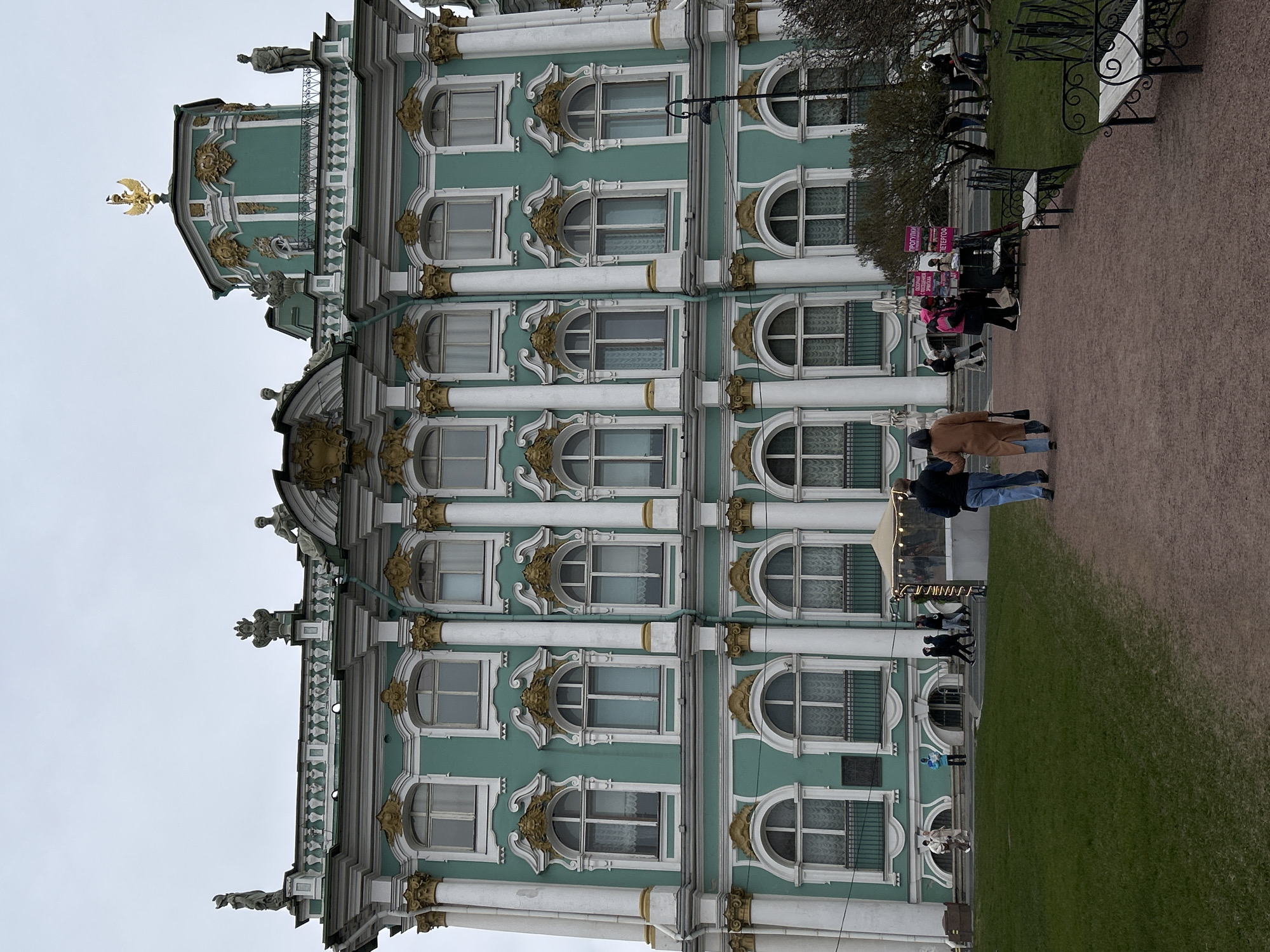 An outdoor scene captures a grand, ornate building under an overcast sky, characteristic of the Winter Palace or Hermitage Museum in Saint Petersburg, Russia. The building features a distinctive light teal or mint green facade adorned with elaborate white architectural trim, columns, pilasters, and intricate gold decorative elements. Multiple stories are visible, each with numerous windows, a mix of rectangular and arched shapes, framed with white and often topped with golden embellishments.
The roofline is detailed with white balustrades and sculpted figures. A prominent golden double-headed eagle, a symbol of Russia, is perched atop a smaller cupola-like structure near the building's peak.
In the foreground and mid-ground, a wide, unpaved pathway or plaza stretches out, bordered by a strip of green grass near the building. Several individuals and small groups of people are scattered along the pathway. Some appear to be casually strolling, while others are gathered around what seems to be a vendor or information stand. Two pink signs are clearly visible near this group, displaying Russian text including "ПРОГУЛКИ" (Tours/Walks) and "ПЕТЕРГОФ" (Peterhof), indicating the likely presence of tour operators. A wrought-iron bench is partially visible on the right side of the pathway.
The scene suggests a cloudy, cool day, possibly in spring or autumn, with soft, diffused light. The overall atmosphere is tranquil, portraying a typical day at a popular historical and tourist attraction.