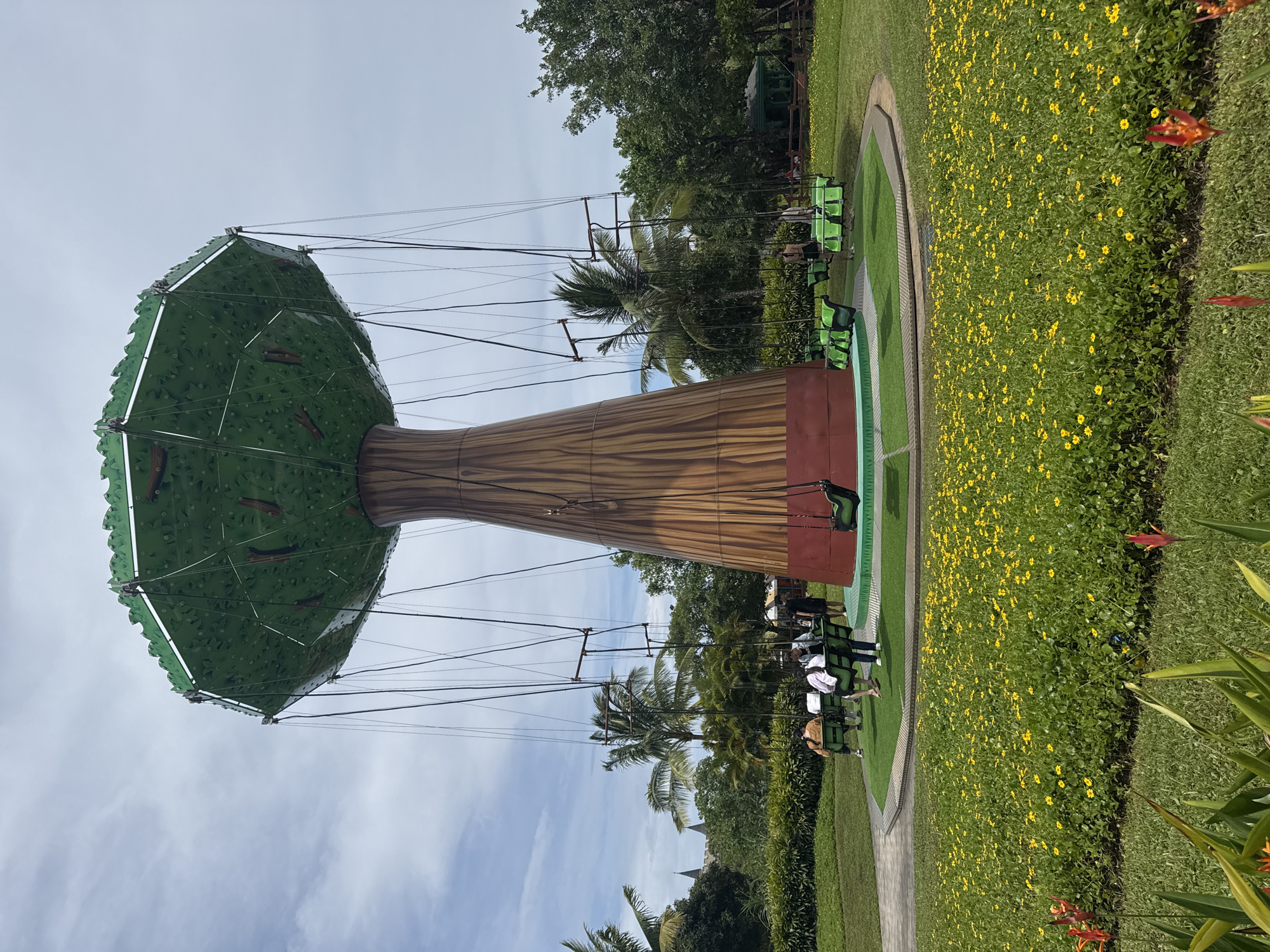 The image shows a children's swing ride, designed to resemble a tree, located in a park-like setting. The "tree" consists of a tall, wooden-looking trunk with a green, leafy canopy overhead. Seats suspended from the canopy by cables allow riders to swing around the structure. People, including children and adults, are seated in the swings. The ride is surrounded by a manicured lawn, bordered by a flowerbed filled with yellow flowers. In the background, various trees and vegetation are visible, along with a blue sky with scattered clouds. The specific location is the Asia Park in Đà Nẵng, Vietnam.