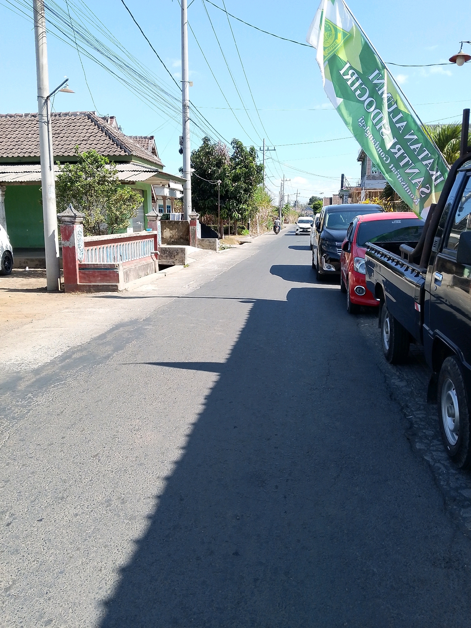 A bright, sunny day in Banjarejo, Indonesia. The image captures a street scene with several vehicles parked or driving along the right side of the asphalt road. The most prominent vehicle in the foreground is a dark-colored pickup truck, partially visible on the far right. Next to it is a red car, followed by a black minivan, and further down the road, a white car and a motorcycle are visible. A large green and white banner is attached to a pole on the right side of the road, angled towards the sky, with text that appears to be in Indonesian. The left side of the street features a green-painted building with a tiled roof and a decorative red and white fence. Trees and utility poles with numerous wires are scattered throughout the scene. The shadow of the vehicles and other objects falls across the road, indicating the direction of the sun.