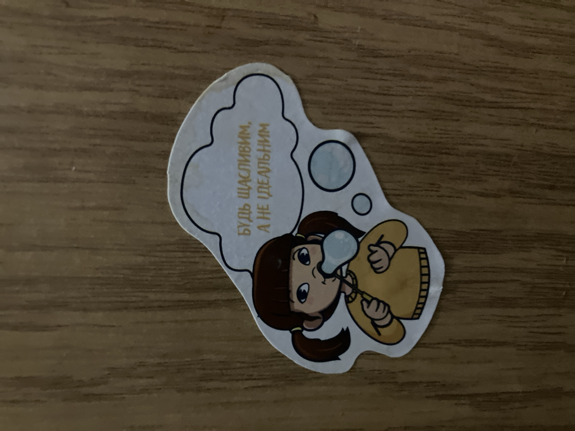 This is a sticker featuring a cartoon illustration of a young girl blowing bubbles. The girl has brown hair styled in pigtails, large blue eyes, and is wearing a yellow sweater. She is shown in profile, blowing a light blue bubble from a bubble wand. Above her head, a thought bubble contains text in Russian.
The text reads: "БУДЬ СЧАСТЛИВЫМ, А НЕ ИДЕАЛЬНЫМ" which translates to "BE HAPPY, NOT PERFECT."
The sticker itself is irregularly shaped, with a scalloped outline for the thought bubble and a more fluid shape for the girl's figure. It is placed on a wooden surface, likely a table or desk, which has a visible wood grain texture. The lighting suggests an indoor setting, possibly during the daytime. There are no specific environmental cues indicating the exact location, but the presence of the Russian text and the provided context of Kyiv, Ukraine, suggest it could be found in that region. The overall tone of the sticker is cheerful and encouraging, with the image of the girl blowing bubbles evoking a sense of innocence and joy.