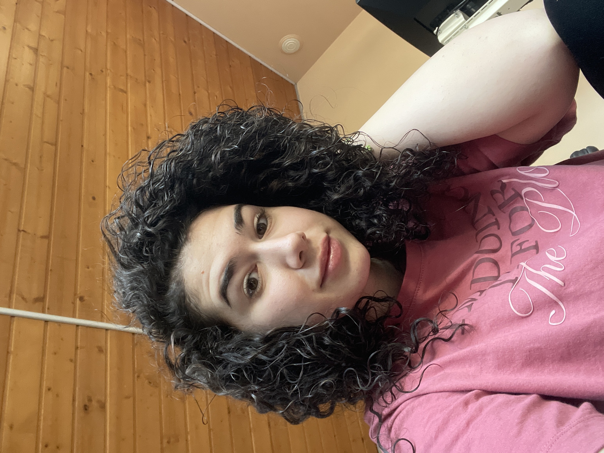 A woman with curly black hair is lying down and looking at the camera. She is wearing a pink t-shirt with white writing. The background consists of a wooden paneled wall and a beige ceiling. Part of a black object is visible in the upper right corner. The location is not discernible from the image, and there is no basis to say that this image was taken in Sofia, Bulgaria.