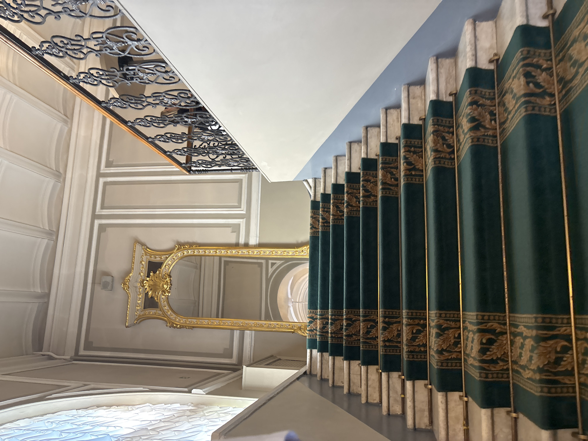 The image shows a grand staircase in Saint Petersburg, Russia, likely within a historic palace or museum. The staircase is carpeted with a deep green runner featuring an ornate gold pattern. The steps themselves are made of white marble. A decorative gilded mirror with elaborate scrolling frames stands at the top of the staircase, reflecting a hallway with an arched ceiling. To the right, a black wrought-iron balustrade with intricate scrollwork lines the staircase, supported by a light blue and white wall. The ceiling above features decorative molding and recessed panels. The overall impression is one of opulence and historical grandeur.