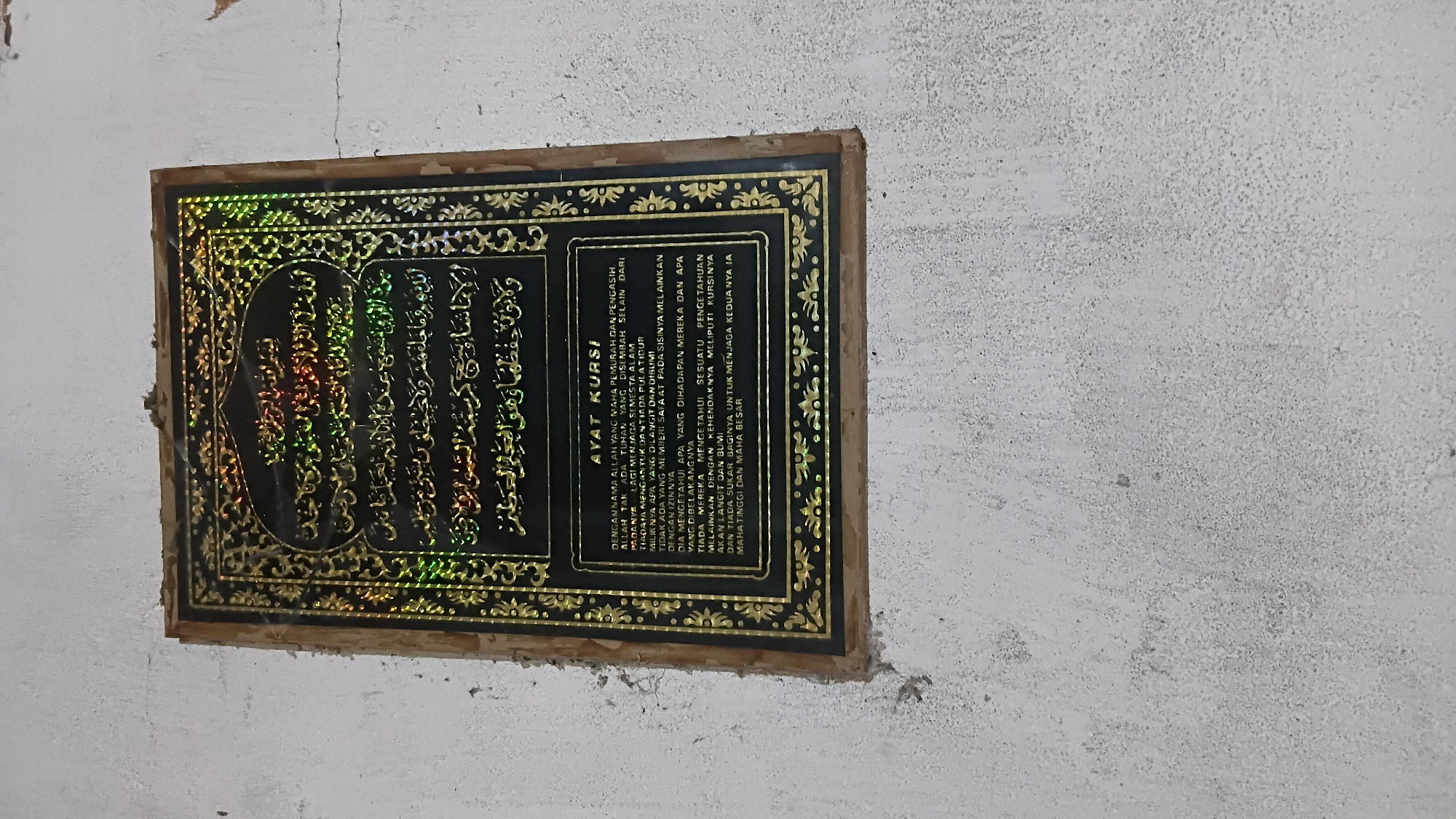 An image shows an Arabic inscription and translation displayed on a wall in Prabumulih, Indonesia. The inscription is presented within an ornate, holographic frame. The text translates to "Ayat Kursi" which is a verse from the Quran. The inscription reads: "With the name of Allah, the Most Gracious, the Most Merciful. Allah! There is no god but He,- the Living, the Self-subsisting, Eternal. No slumber can seize Him nor sleep. To Him belongs whatever is in the heavens and on earth. Who is that that can intercede with Him except with His permission? He knows what is before them and what is behind them, and they shall not comprehend aught of His knowledge but what He wills. His Throne extends over the heavens and the earth, and He feels no fatigue in guarding them. And He is the Most High, the Most Great." The wall appears to be made of concrete and shows signs of age and wear.