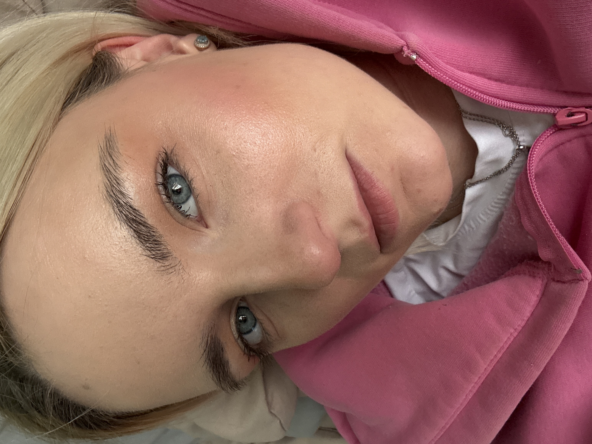 A close-up portrait shows a light-skinned woman with blonde hair, blue eyes, and visible makeup, including blush. She is wearing a pink hoodie and a white shirt with a silver necklace underneath. The location in the city of Vsevolozhsk, Russia is not discernable from the image.