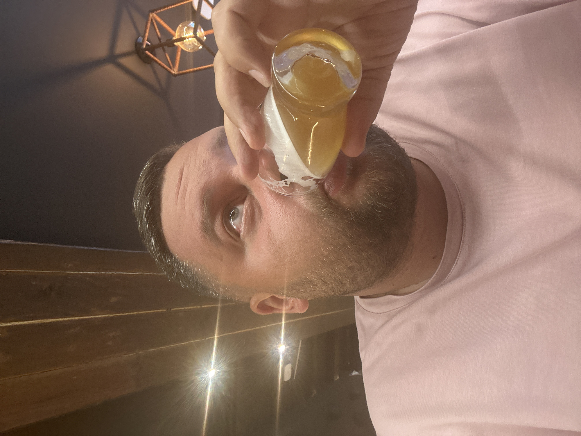 An indoor scene, rotated 90 degrees counter-clockwise, depicts a man consuming a beverage in Encamp, Andorra. The primary subject is a man, shown from the nose upwards, with short dark hair displaying some grey, and a beard. He is wearing a light pink t-shirt. His head is tilted back as he drinks from a clear shot glass, which his hand holds. The glass contains a yellow or amber liquid, some of which appears to be spilling or dripping near his chin. His eye is open and looks towards the viewer.
The background reveals a dark ceiling with a geometric, open-cage style light fixture containing an illuminated Edison bulb. Below this, horizontal wooden panels are visible, possibly part of a wall or furniture. The lower portion of the image shows a darker surface with bright, starburst-like reflections from light sources, suggesting a reflective tabletop or bar. The artificial lighting indicates an evening or nighttime setting. There is no visible text in the image.