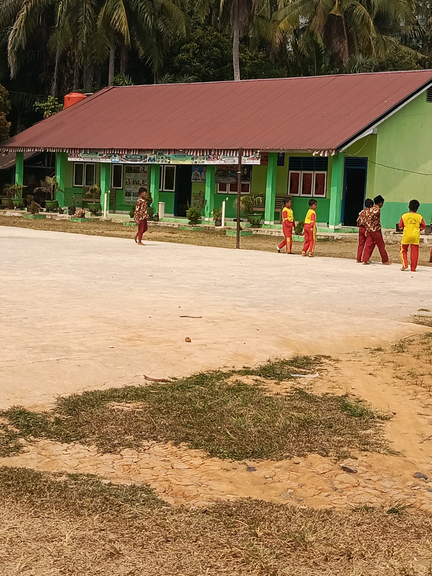 In the city of Megang Sakti IV, Indonesia, a light-colored concrete schoolyard features several children in uniforms. A light green, single-story school building with a red metal roof is visible in the background. The building has multiple windows with red frames and a blue door. In front of the building, potted plants are arranged along a covered walkway. Several children are walking across the schoolyard. One child, wearing a patterned shirt and red pants, walks alone towards the left. Further to the right, four children in yellow shirts and red pants are walking together. The schoolyard is bordered by patches of dry grass and some trees in the distance.