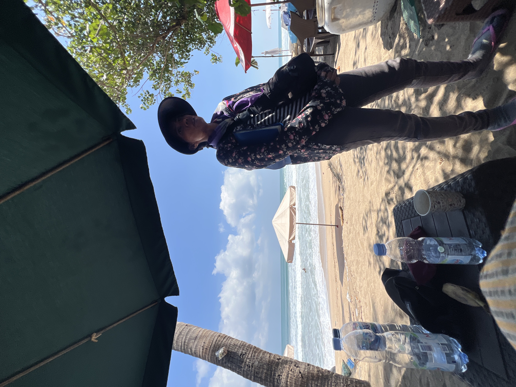 A person stands on a sandy beach in Kuta, Indonesia, under a dark green umbrella. The person is wearing a dark hat, a patterned long-sleeved shirt, and dark pants. They are looking towards the ocean. In the foreground, there are two plastic water bottles and a black bag on a dark table. In the background, the ocean has waves, and there are beach umbrellas. A palm tree is visible to the left. The sky is blue with some clouds.