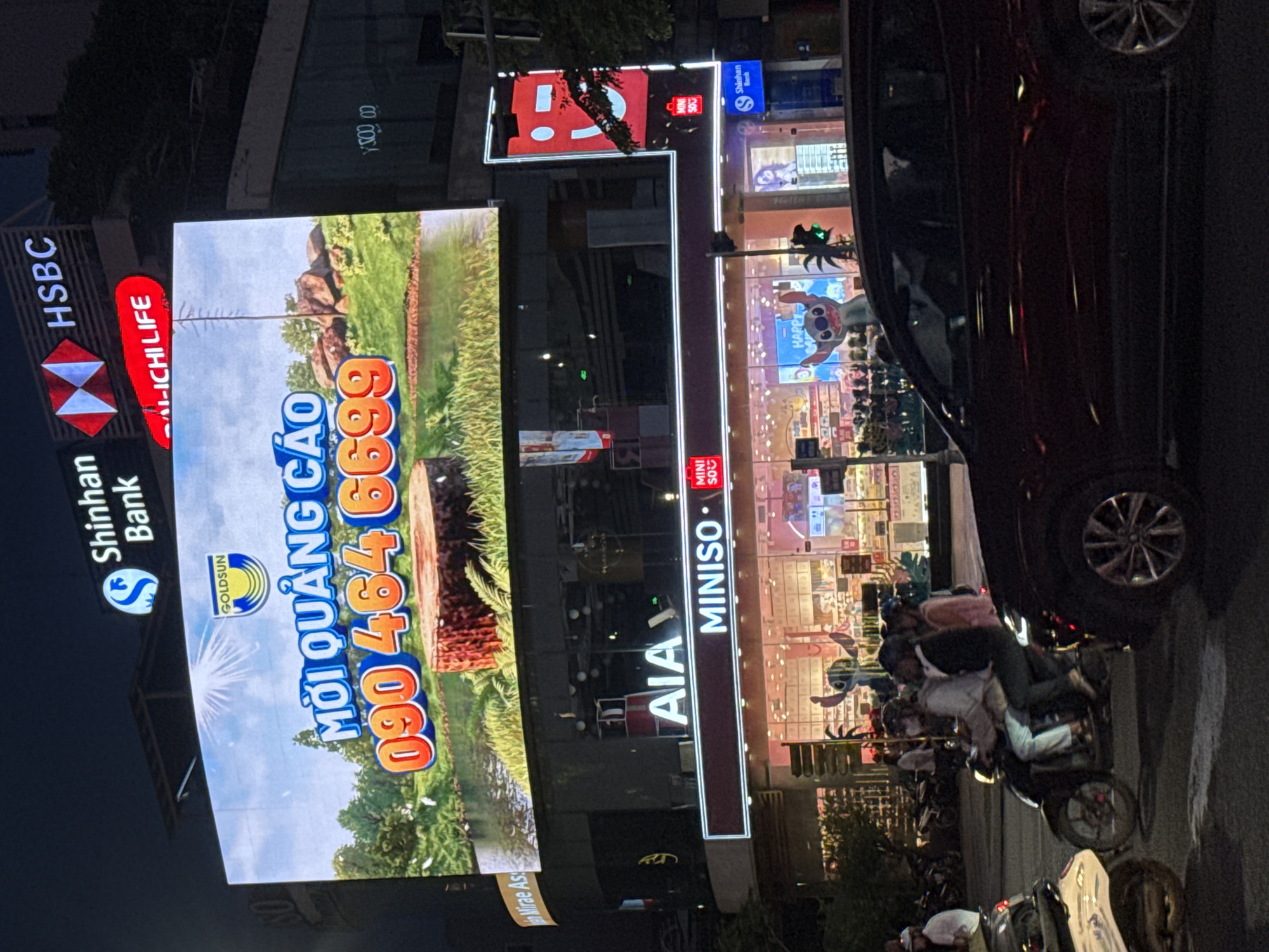 Captured at night, the image reveals a bustling street scene in Đà Nẵng, Vietnam. Dominating the view are several illuminated billboards. One advertises 'AI-ICHI LIFE' and the number '090 464 6699.' Another displays the Shinhan Bank logo and the HSBC logo. A third announces the presence of MINISO and AIA. A dark vehicle occupies the right side of the frame, partly obscuring the store behind it. On the street, a group of individuals are riding motorbikes.