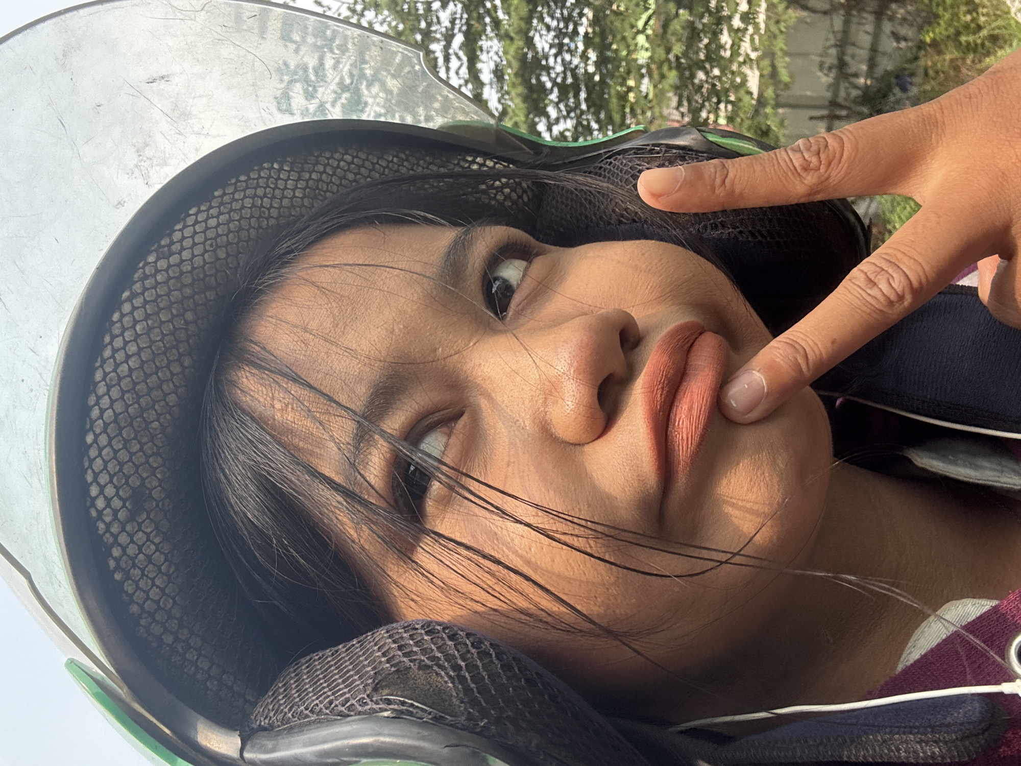 A close-up selfie shows a young woman wearing a green and black helmet with a clear visor, obscuring the top of her head. Her dark hair is slightly messy, with strands falling across her face. She is looking upwards and to her right, with her lips pursed and her right index finger touching her chin. Her right hand is raised, forming a peace sign with her index and middle fingers extended. The background consists of blurred green trees and foliage, suggesting an outdoor, possibly urban, environment. The lighting indicates it is daytime. The subject appears to be on a scooter or motorcycle, given the helmet and typical use of such transportation in cities like North Jakarta, Indonesia.