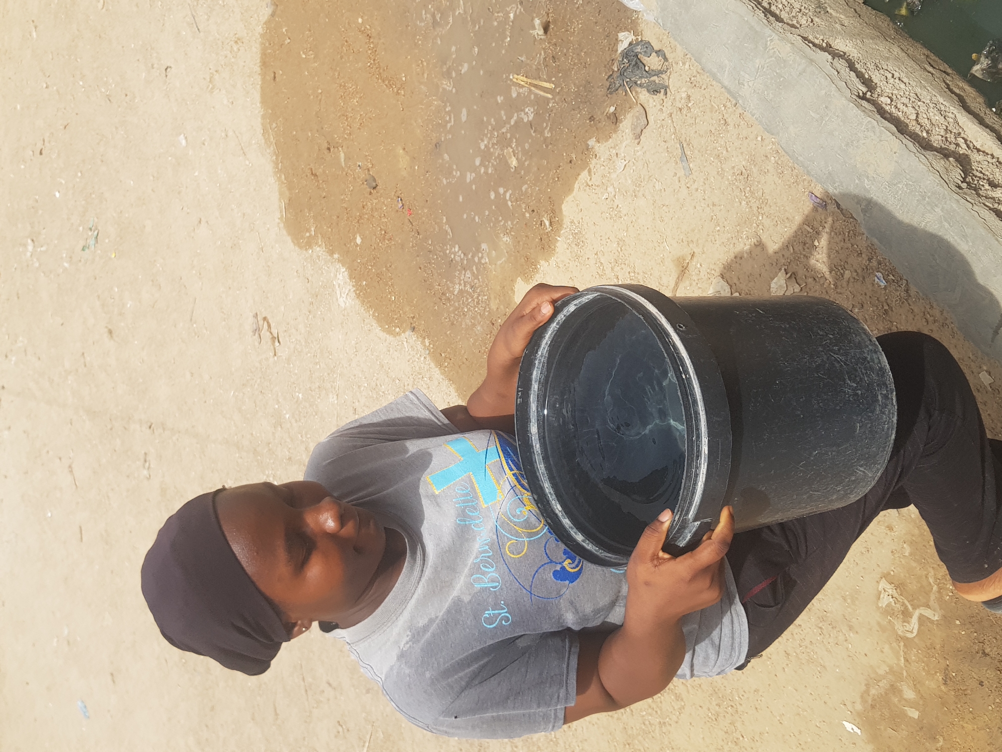 The photograph depicts a woman holding a large, black, cylindrical bucket filled with water. She is wearing a gray t-shirt with blue and gold designs, dark-colored leggings, and a dark head covering. The woman's posture indicates she is holding the bucket firmly. The environment appears to be an outdoor space with a combination of concrete, sand, and dirt surfaces. There's evidence of debris, including what looks like trash, scattered around. The location is indicated as Dutsinma A, Nigeria.